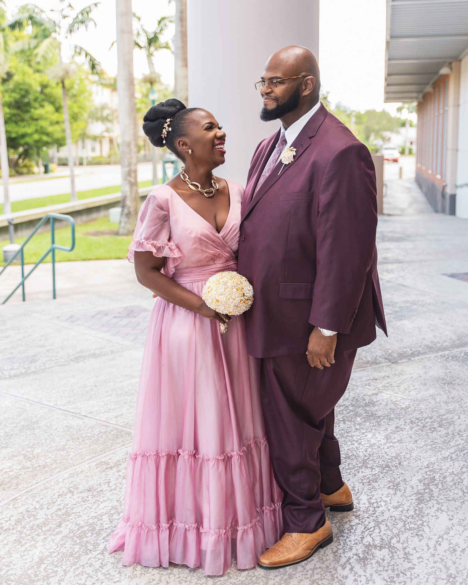 A couple poses outdoors, with her in a pink ruffle dress and him in a burgundy suit, sharing a romantic moment.