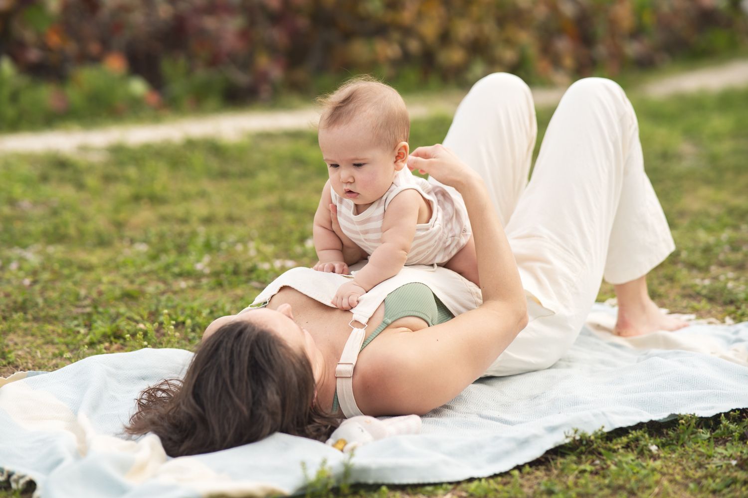 A parent and baby play together while lying on a white blanket in a grassy outdoor setting.