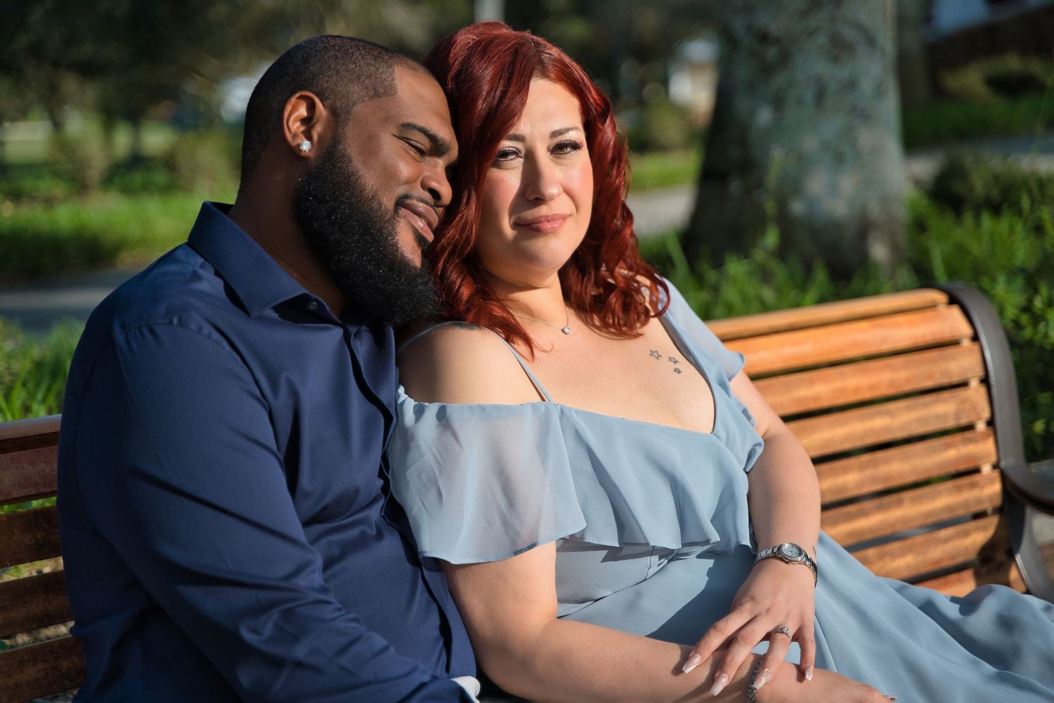 A couple sharing a tender moment while sitting on a wooden bench in a sunny outdoor setting.