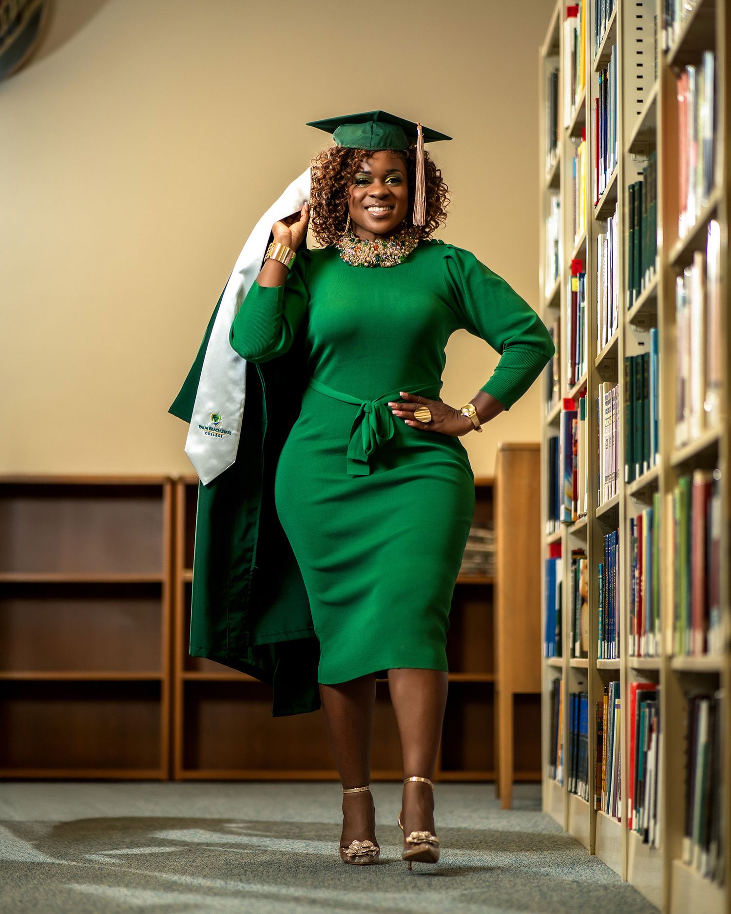 Graduate in emerald green dress and cap poses confidently in library.