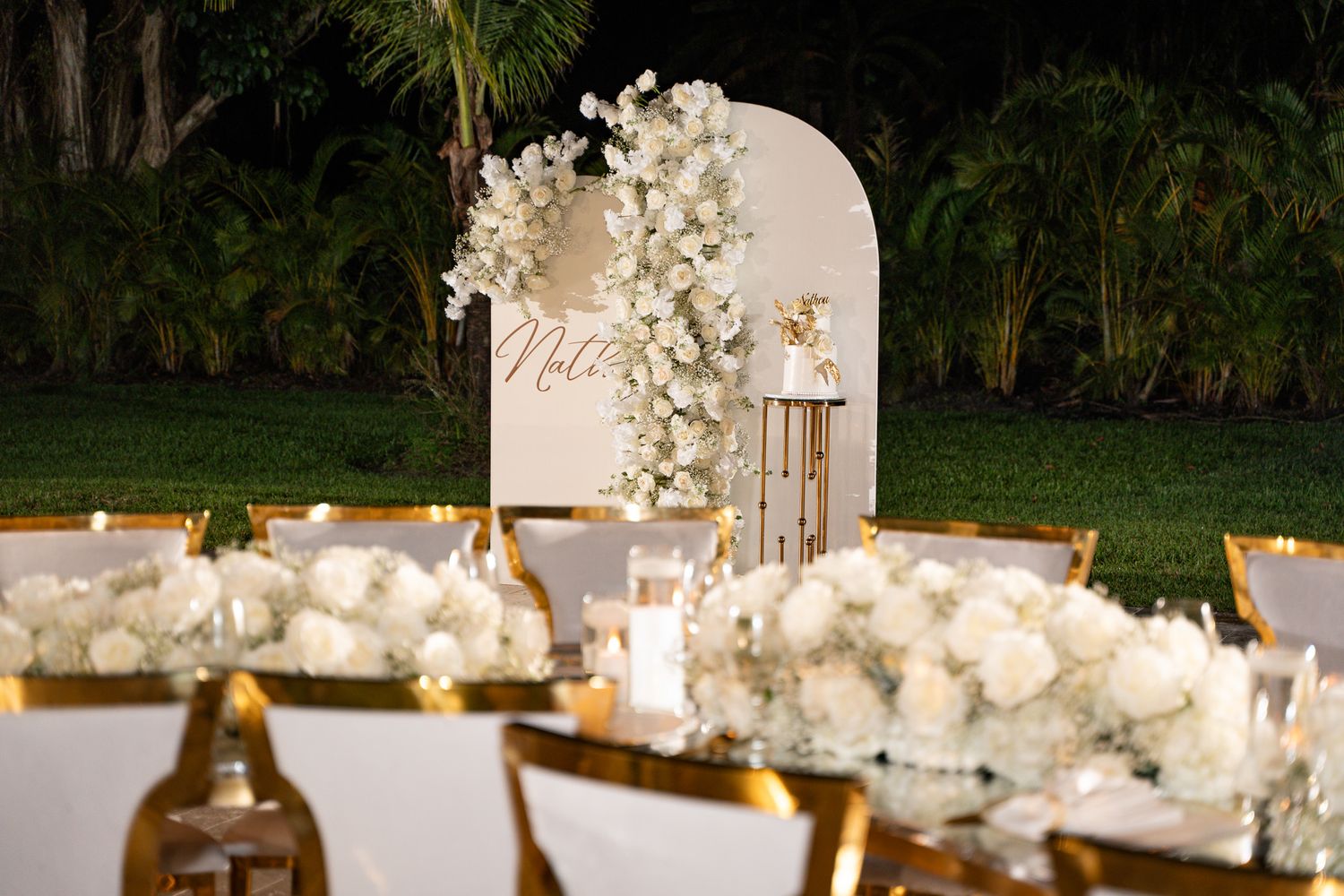 Wedding reception table setup with white floral centerpieces and gold accents against a curved backdrop.