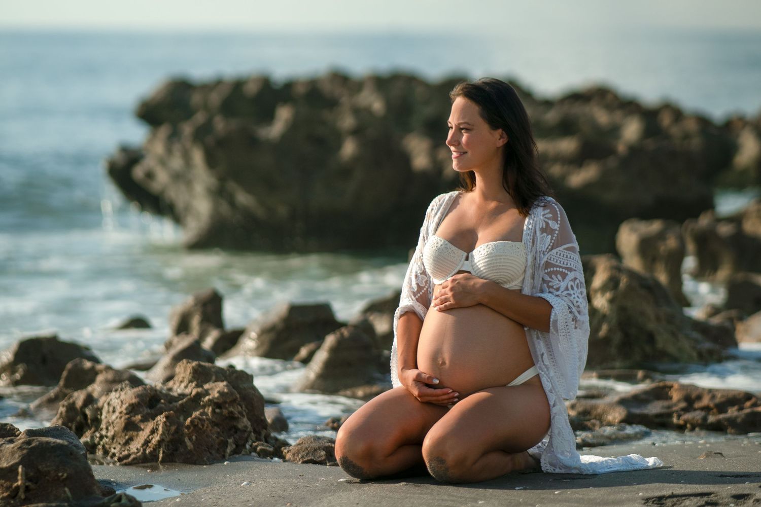 Peaceful maternity moment captured on rocky beach at sunset with ocean backdrop.