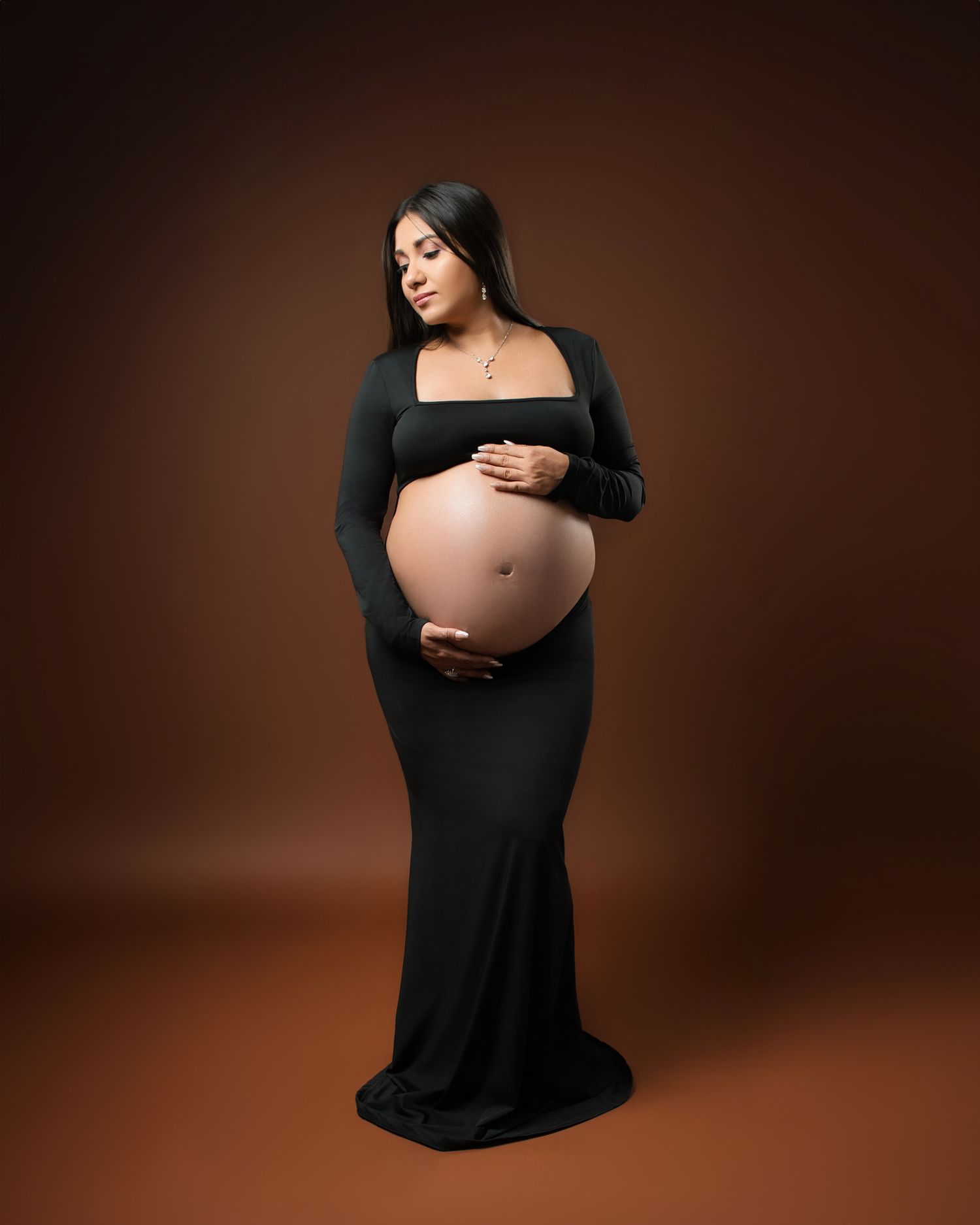 Elegant maternity portrait in fitted black dress against warm brown background.