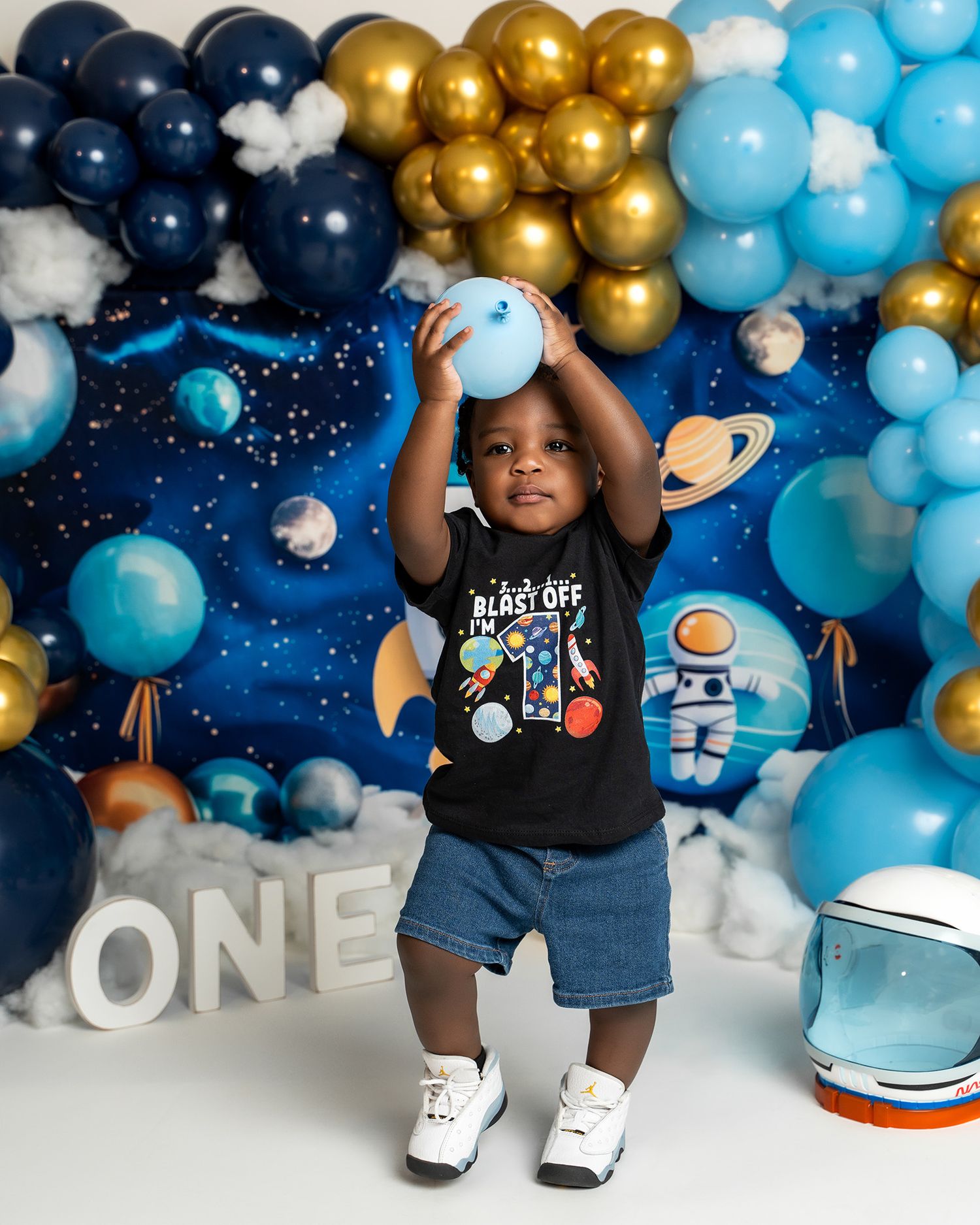 Toddler in space-themed outfit playing with balloons against cosmic party decoration backdrop.