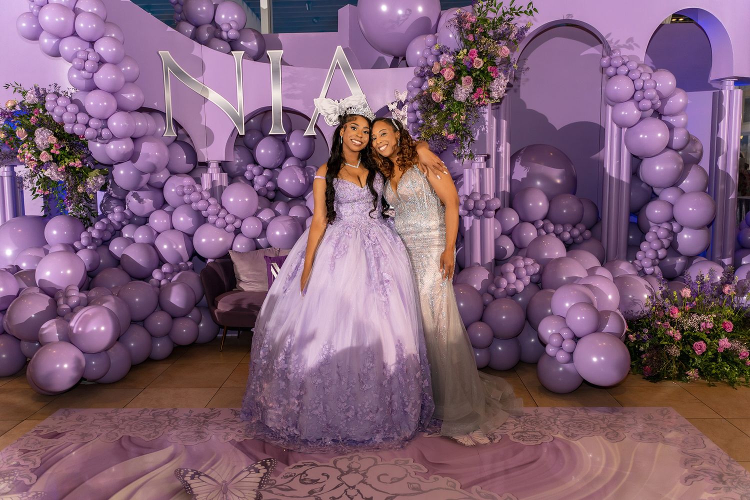 Two people pose in front of an elaborate purple balloon arch and floral display at a formal event.