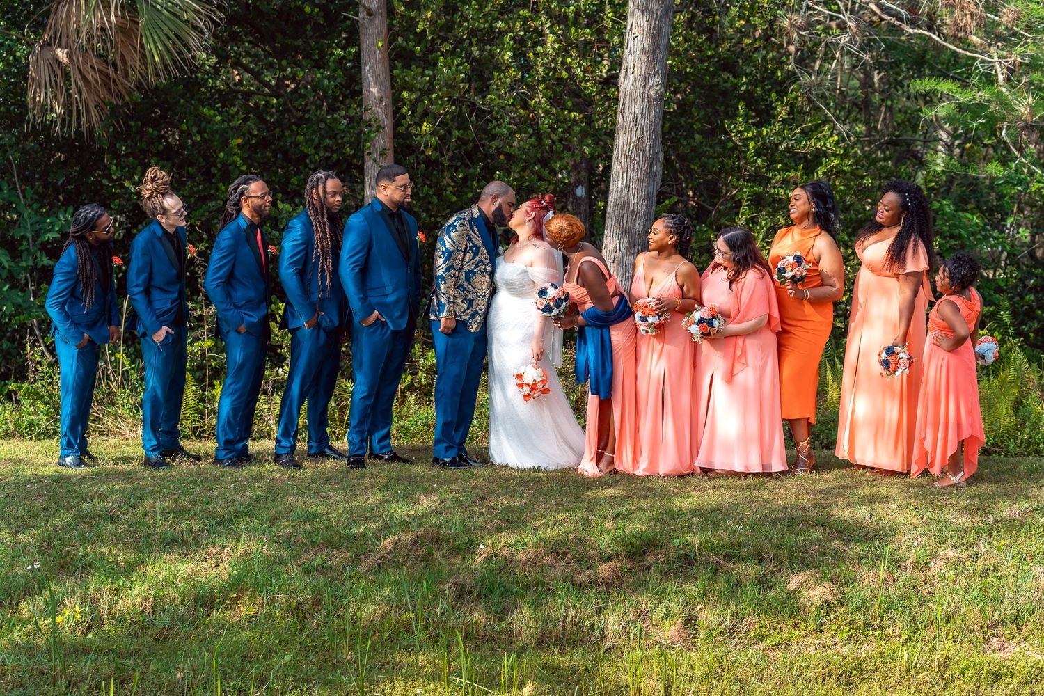 Wedding party lined up outdoors with bridesmaids in coral dresses and groomsmen in royal blue suits.