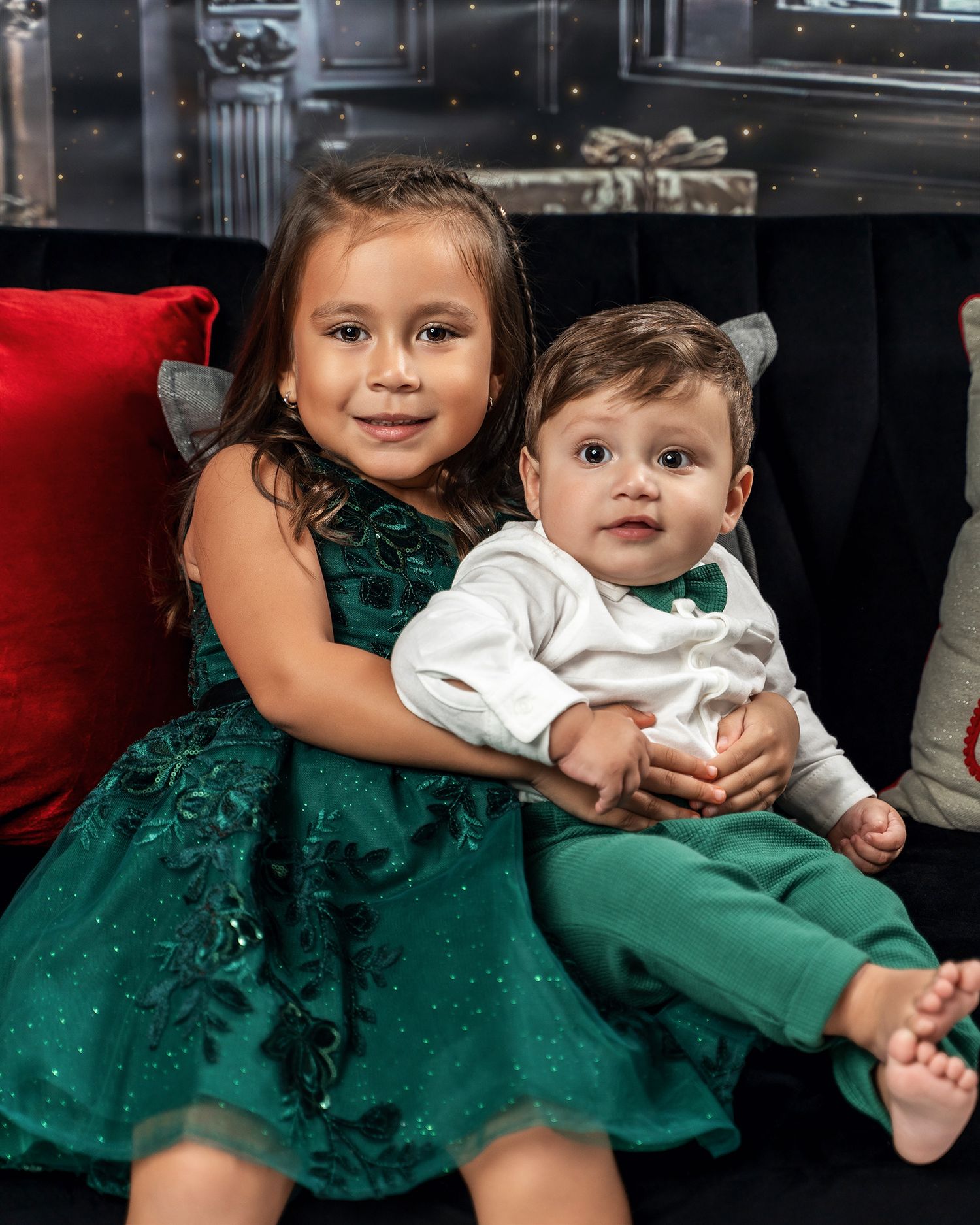 Two children in coordinated green holiday outfits sitting together on decorated Christmas set.