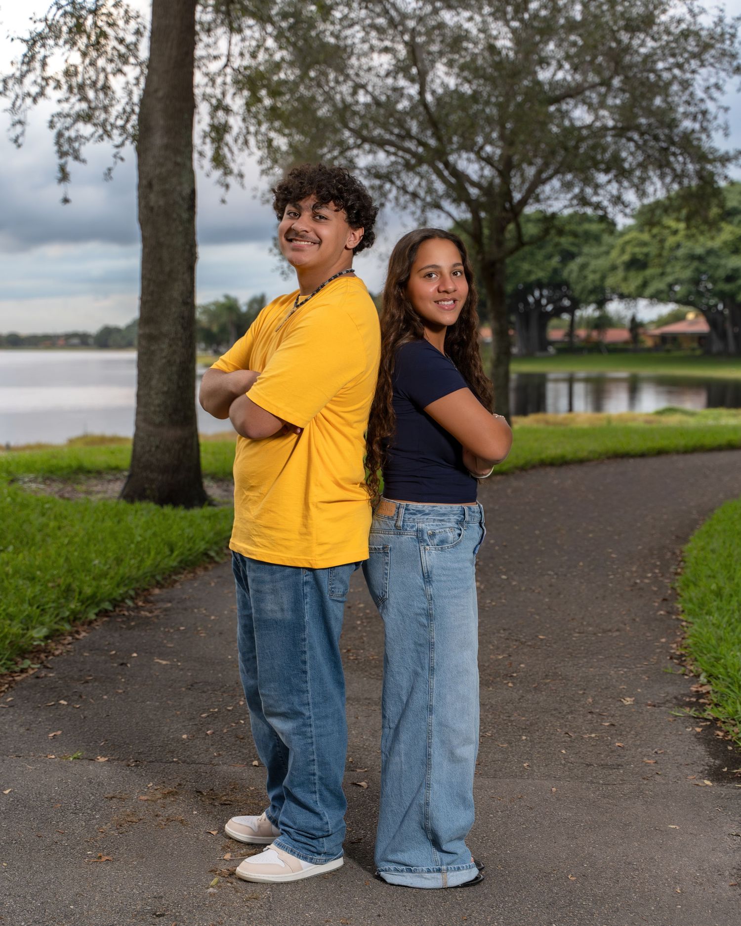 Two people in denim and yellow stand together on a path near trees and water in a playful outdoor photoshoot.