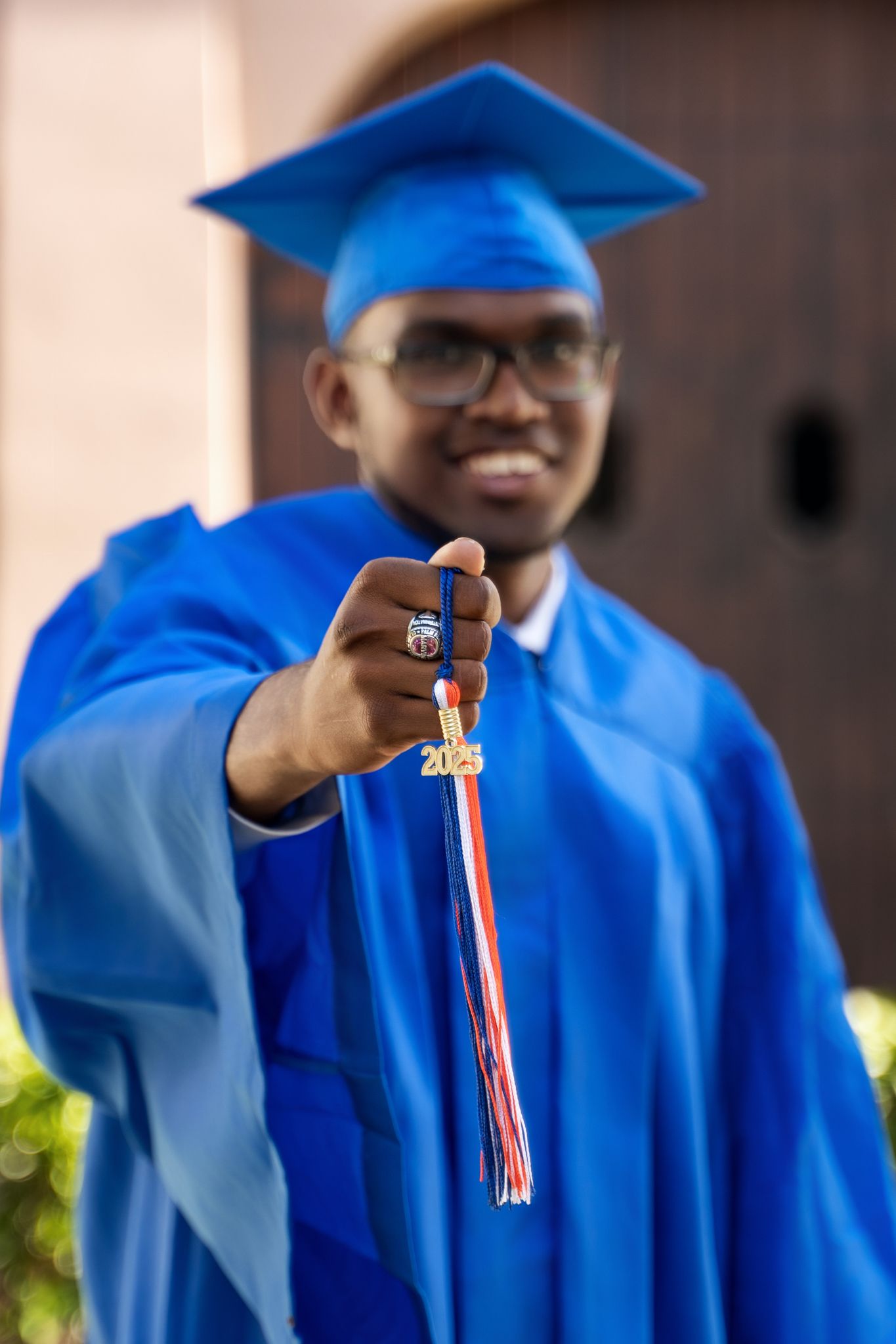 Graduate in blue academic regalia shows off graduation tassel while smiling during ceremony.