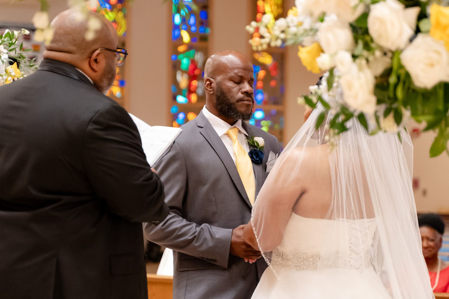 Wedding ceremony in progress with clergy and couple at church altar surrounded by floral arrangements.