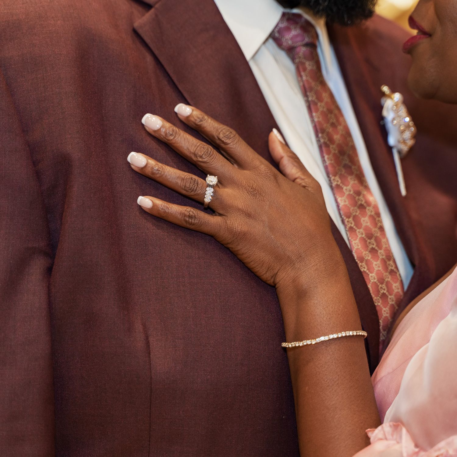 Close-up of hands showing an engagement ring against a burgundy suit jacket.