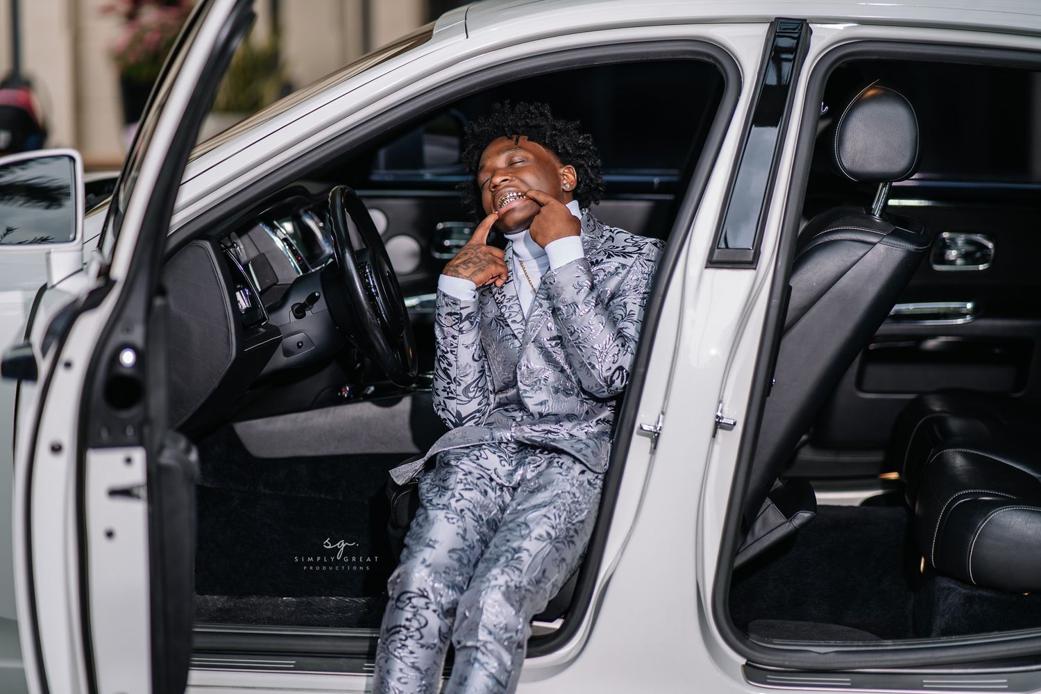 Person wearing gray patterned suit sitting in driver's seat of white car with hand covering face.