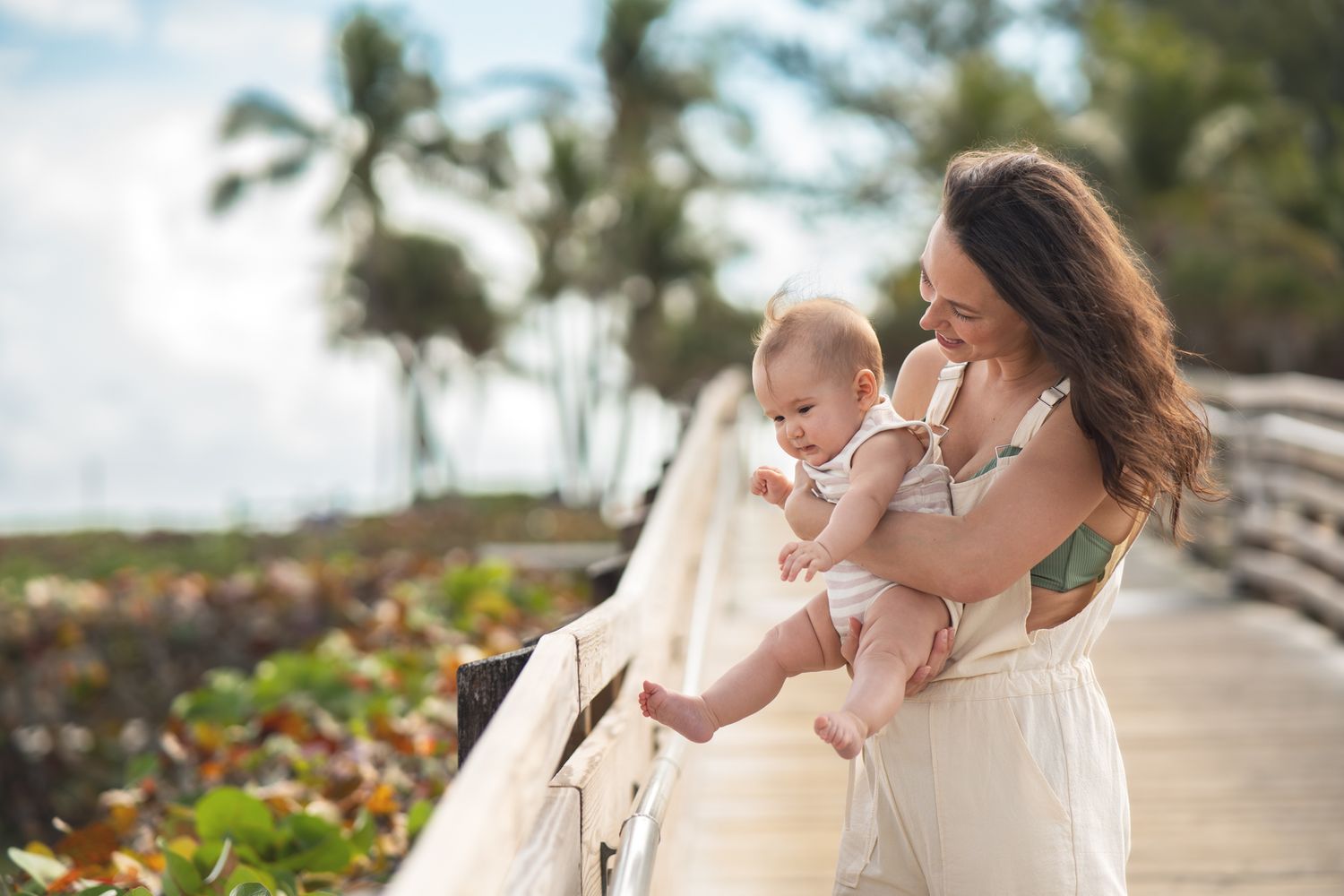 A parent and baby enjoy a sunny moment together on a tropical boardwalk with palm trees.