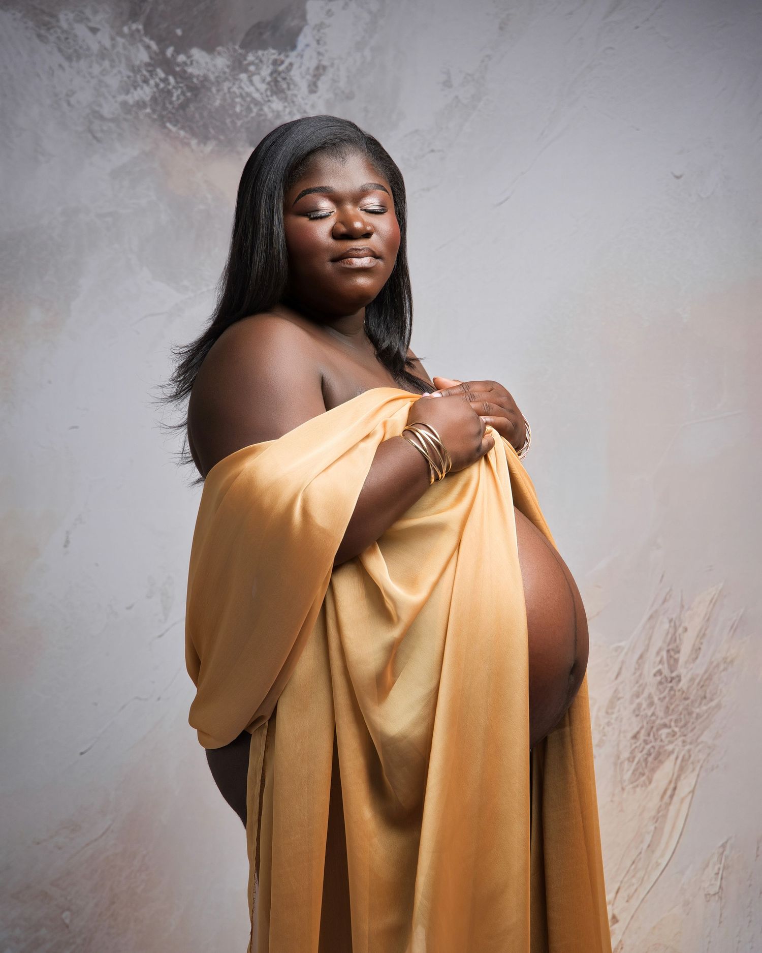 Artistic maternity portrait featuring flowing gold fabric draping against textured grey background.