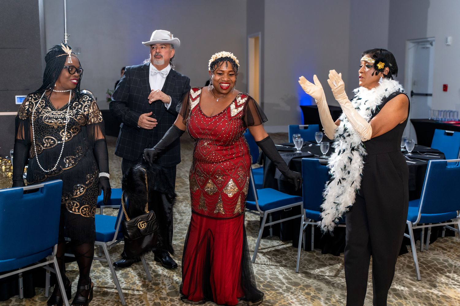 Guests in elegant evening wear dance and celebrate at a formal party with blue chairs in the background.