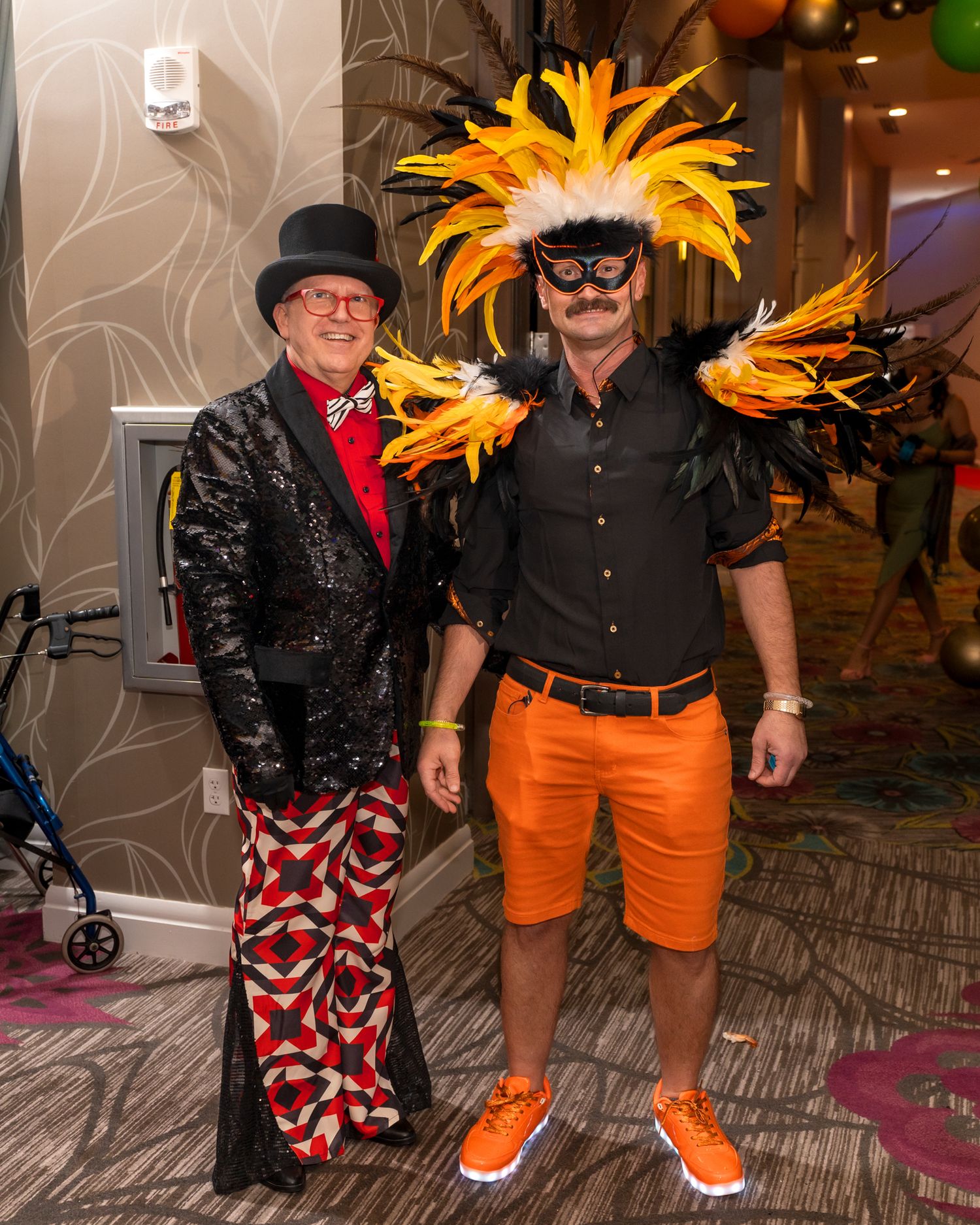 Two people in festive costumes with feathered headpieces posing at a carnival-themed party.
