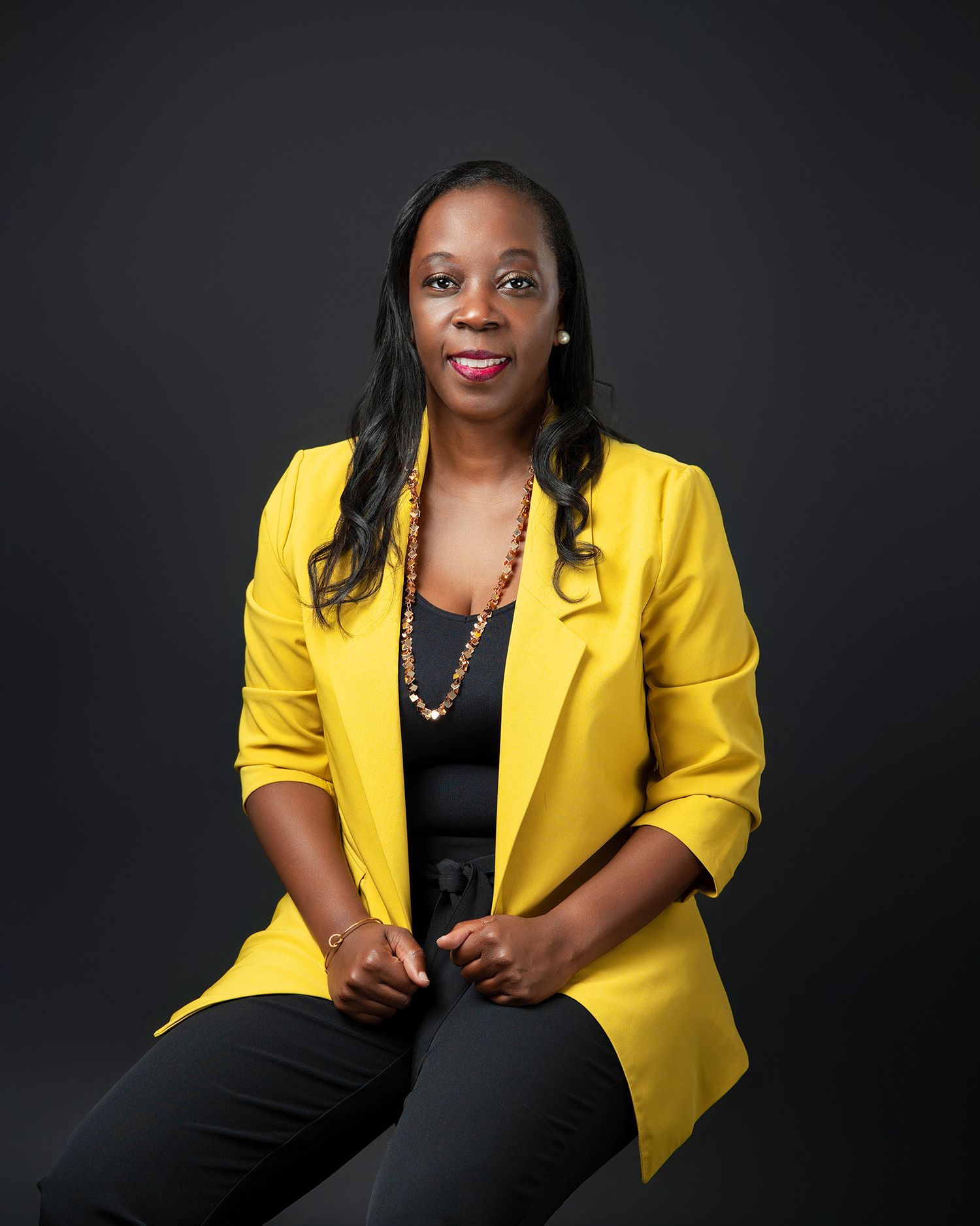 Professional portrait of someone wearing a yellow blazer and necklace against dark background.