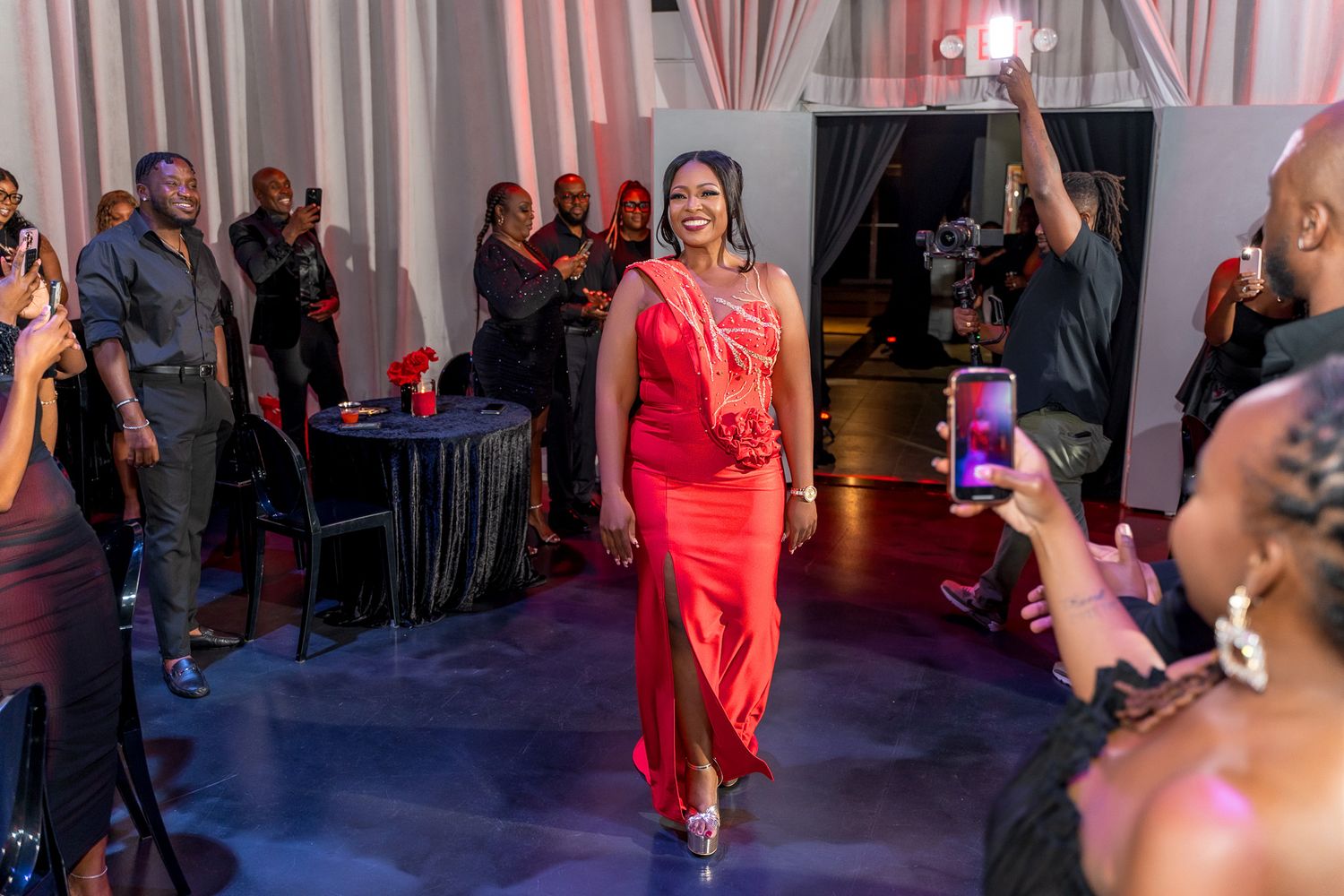 A woman in a striking red dress walks confidently across a stage at an elegant indoor event.