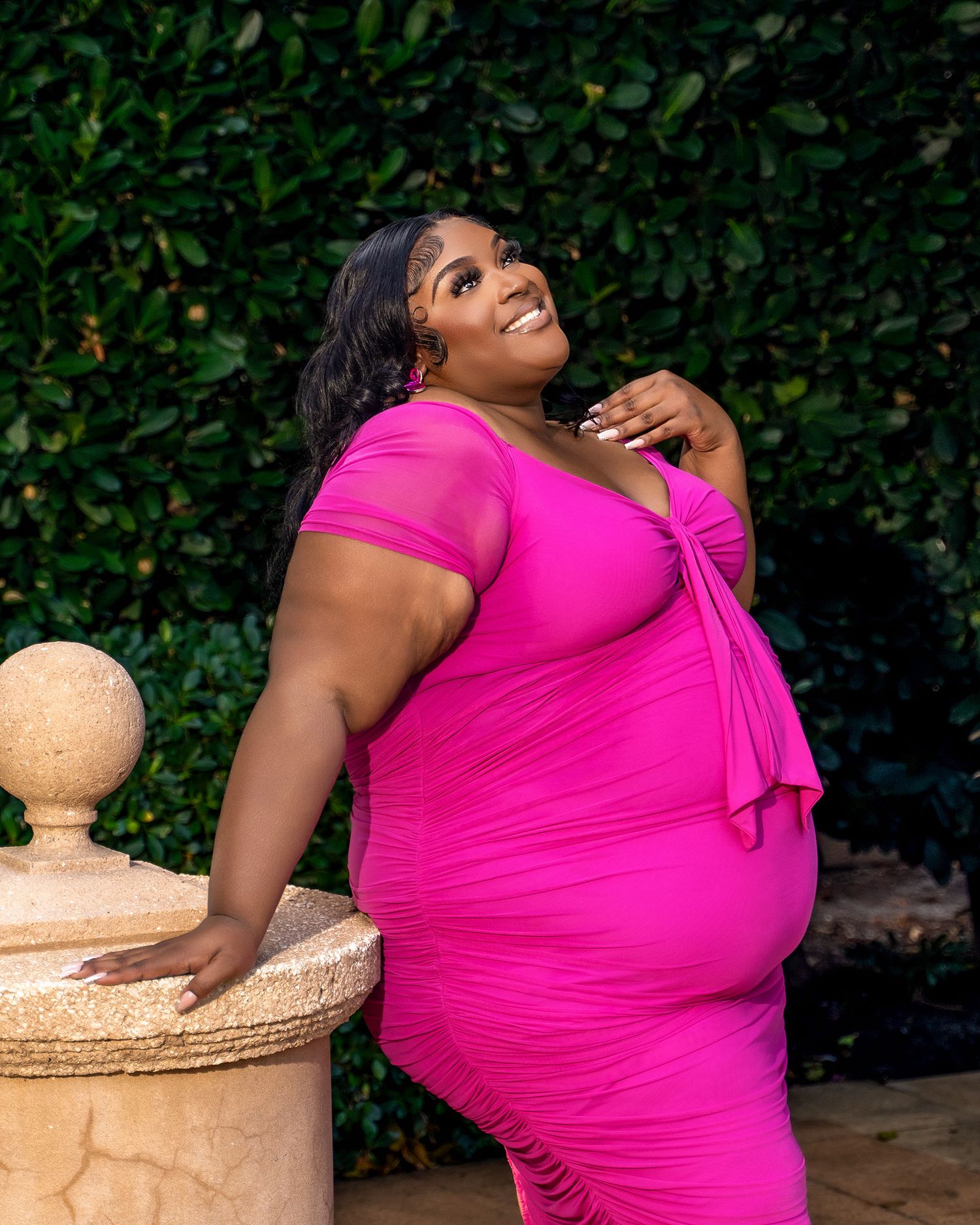 Woman in bright pink dress poses confidently by stone pillar in garden setting.