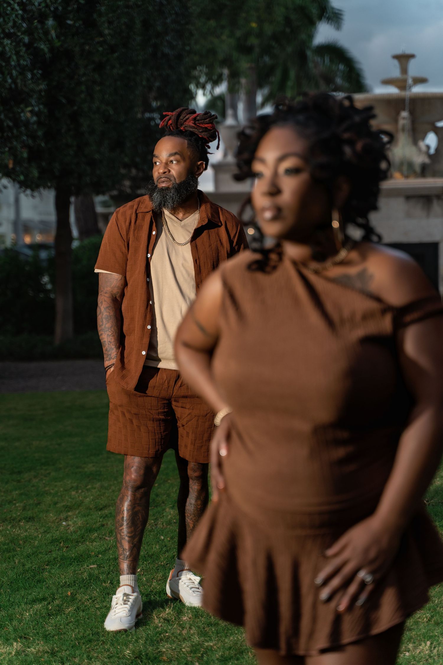 Two people in brown outfits pose outdoors at dusk with palm trees and a fountain in the background.