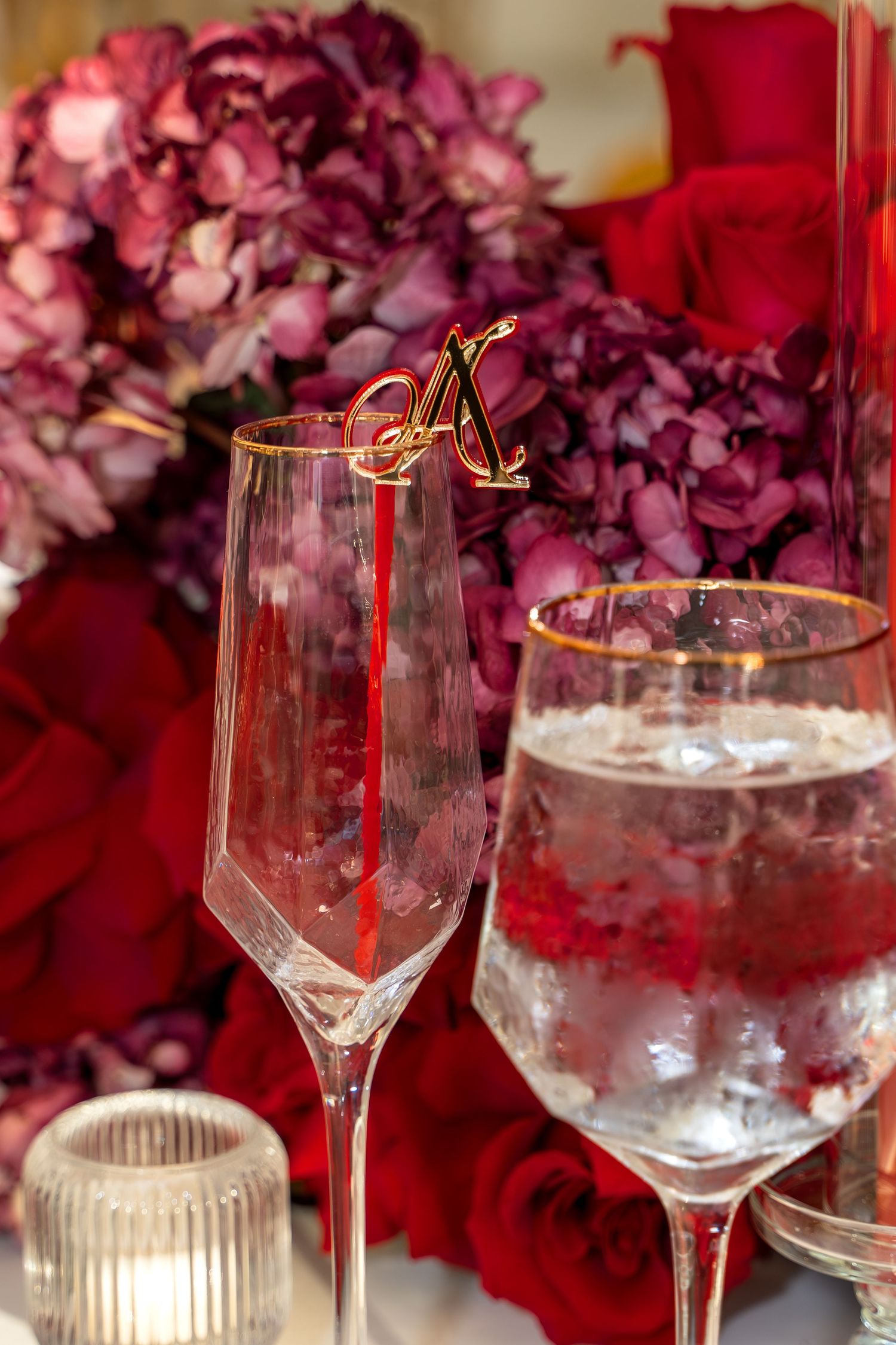 Crystal wine glasses and champagne flutes arranged with deep red floral centerpieces and gold accents.