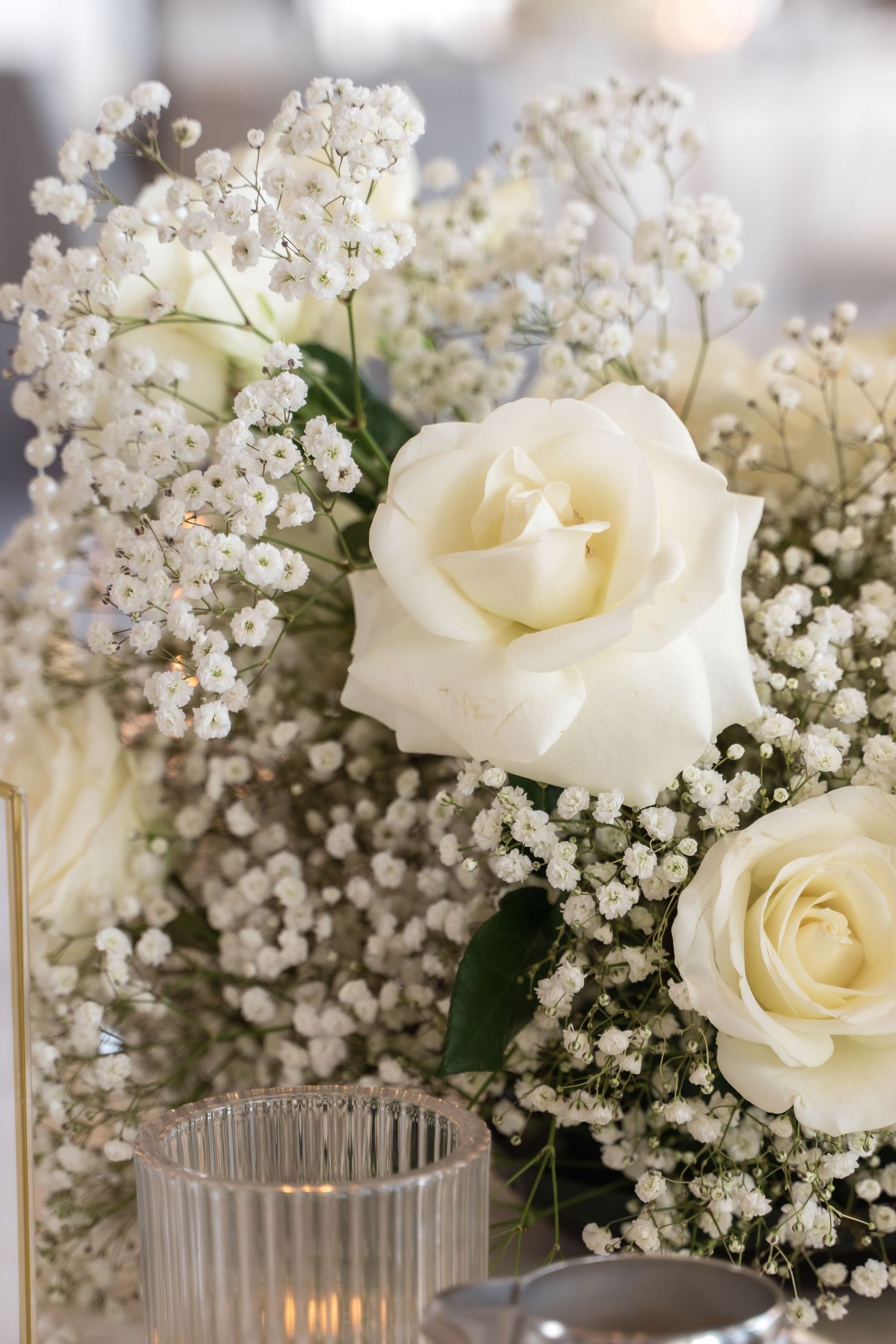 Elegant white roses and baby's breath floral arrangement next to a glass candleholder with soft glowing light.