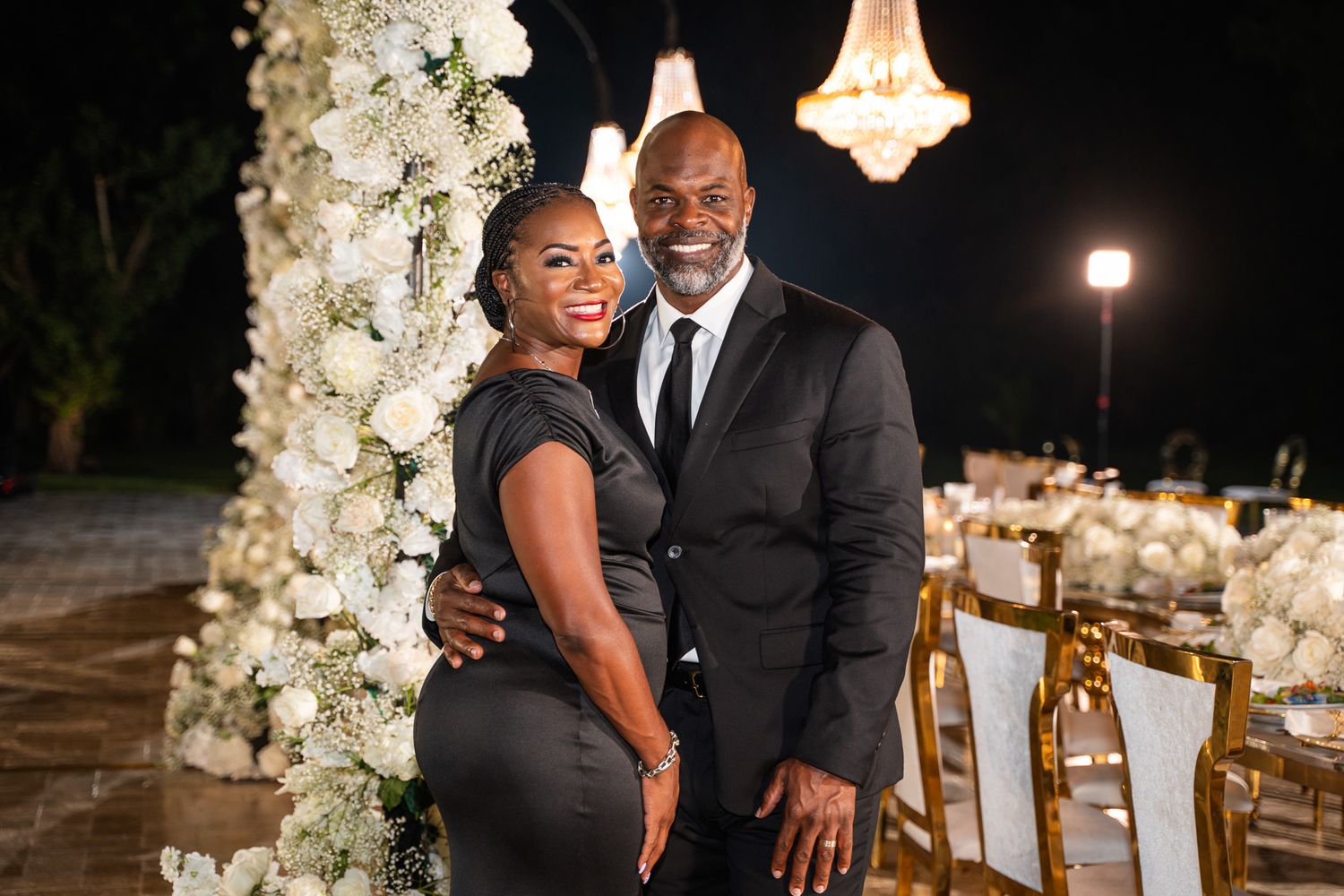 A couple poses in formal attire in front of a white floral arch and chandelier at night.