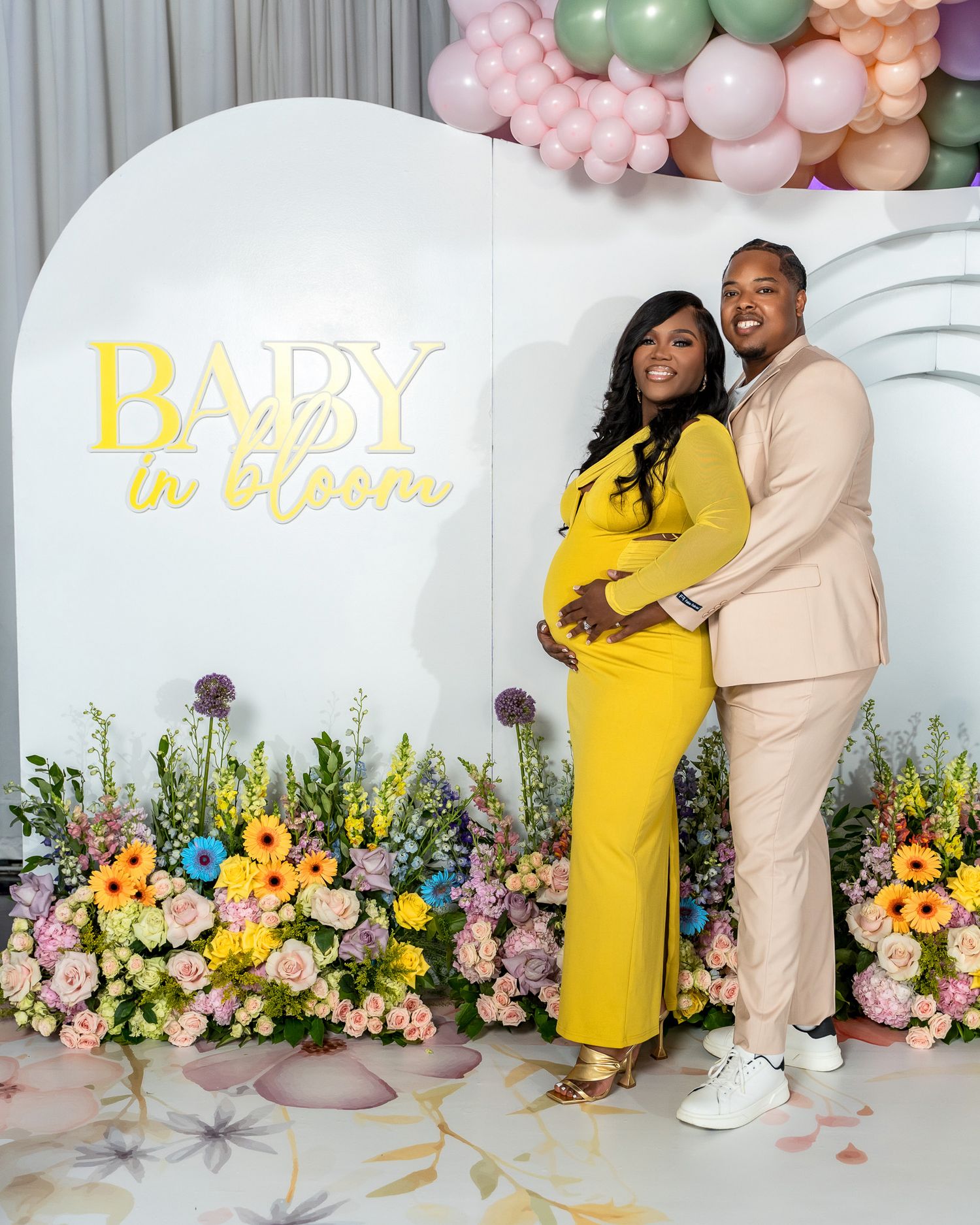 Baby shower celebration setup with floral decor, balloon arch, and 'Baby in bloom' neon sign against white backdrop.