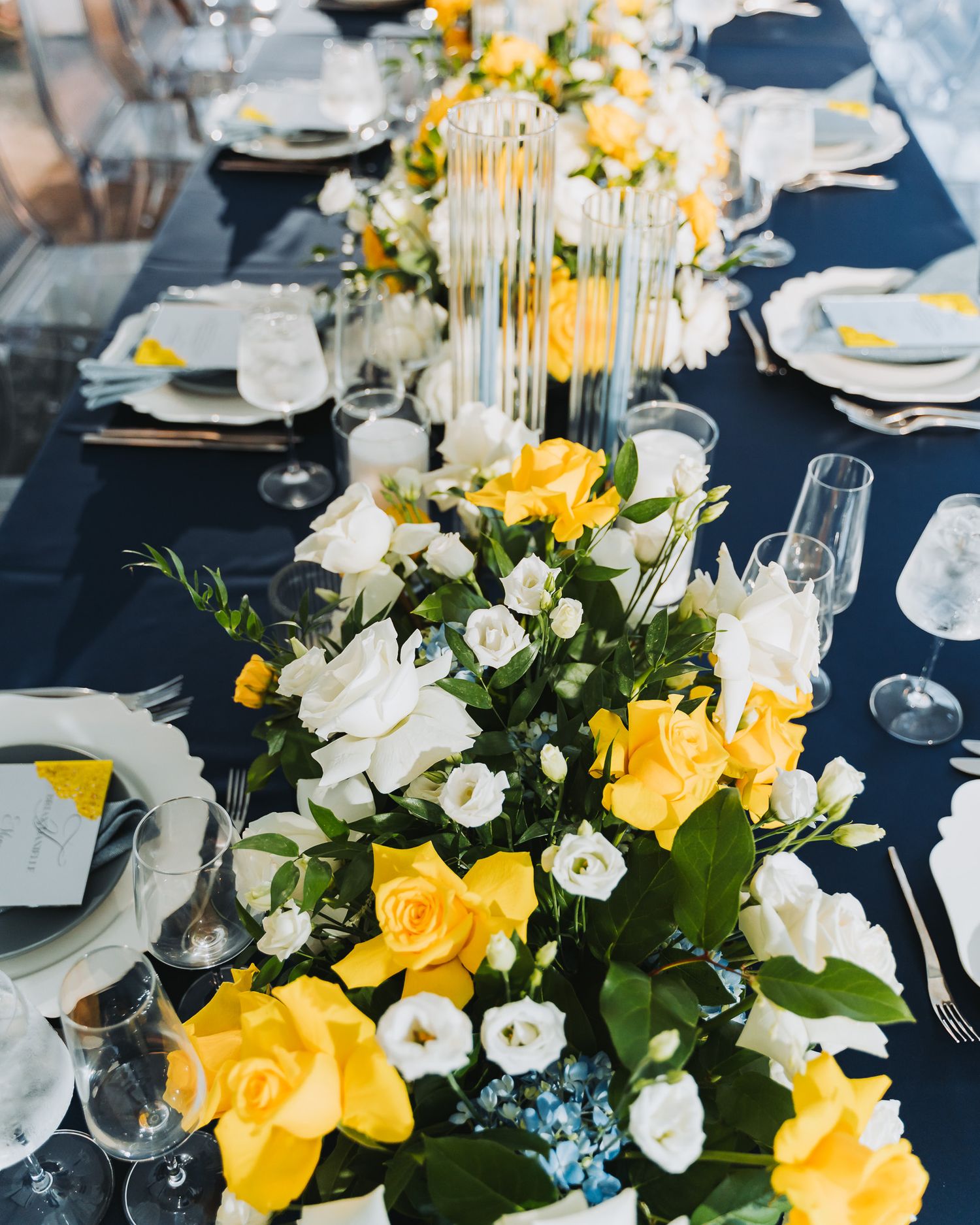 Elegant wedding table setting with yellow and white floral centerpieces on navy tablecloth.