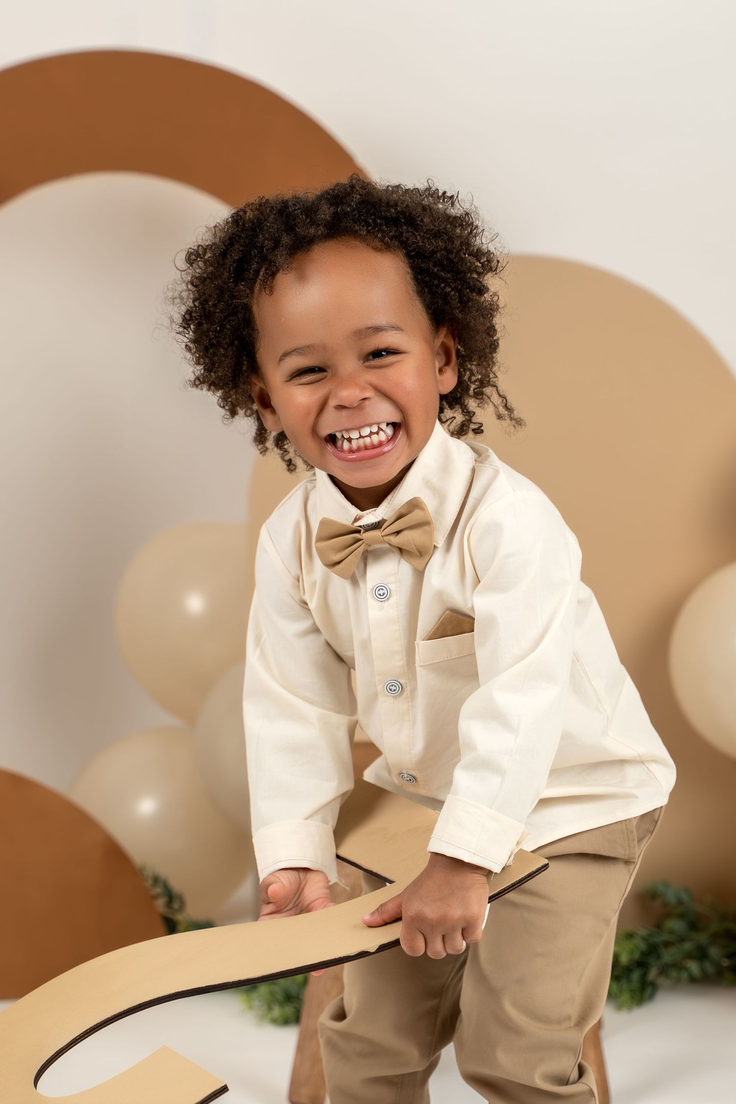 A joyful child in formal cream-colored attire smiles and poses against a brown and white backdrop.