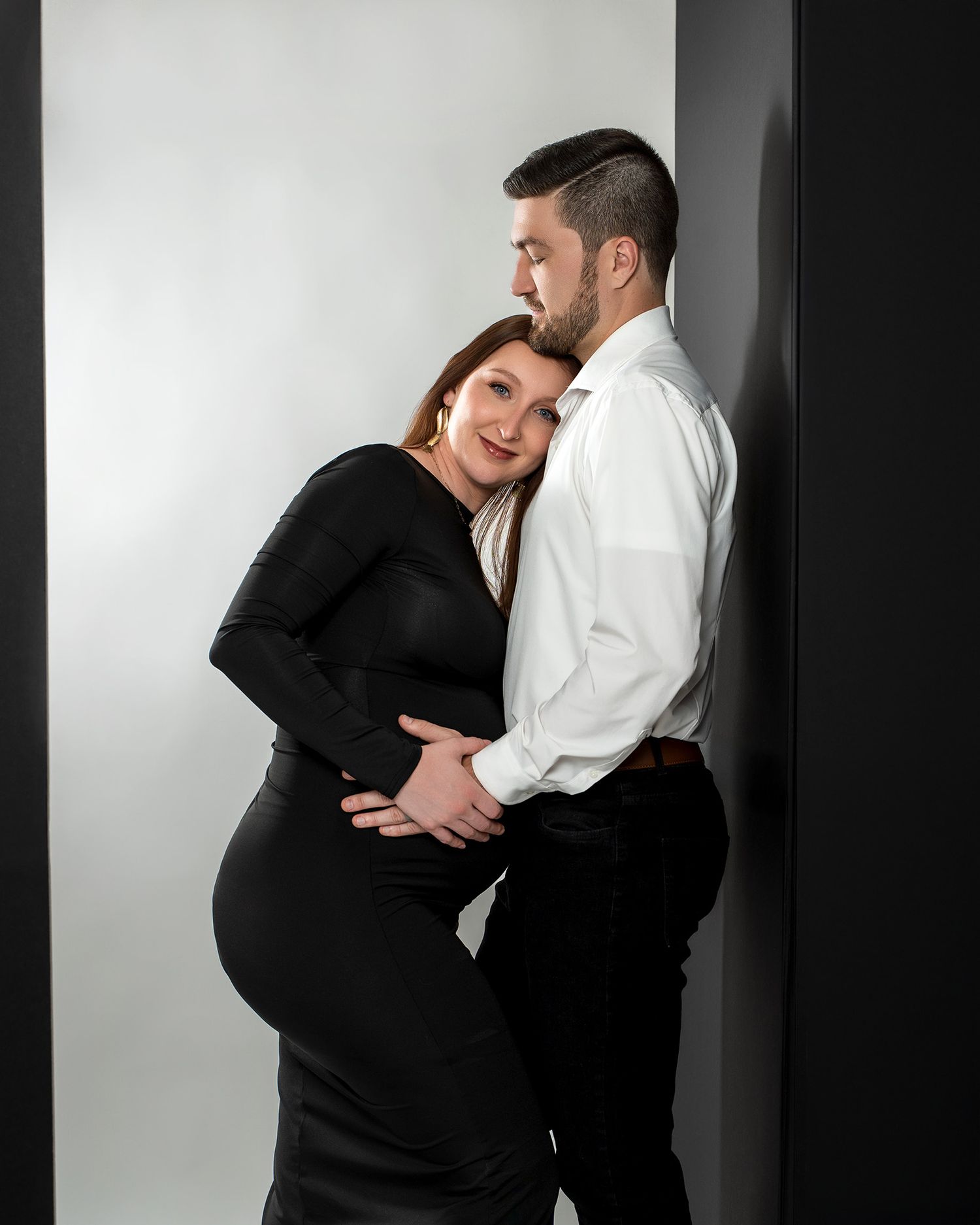 Studio maternity portrait of couple in formal attire embracing against white backdrop.