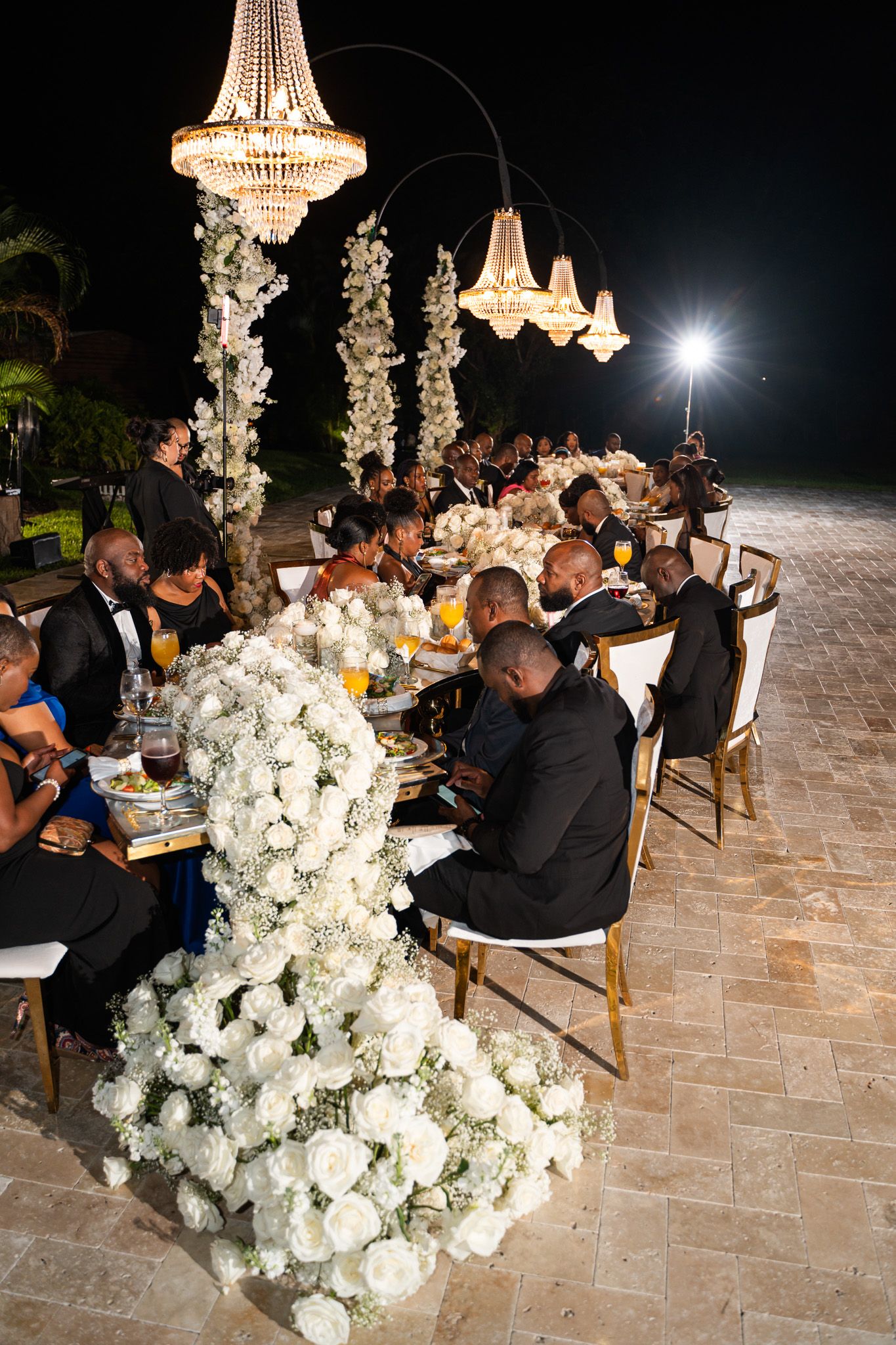 Luxurious nighttime wedding reception with crystal chandeliers and white floral centerpieces on long dining tables.