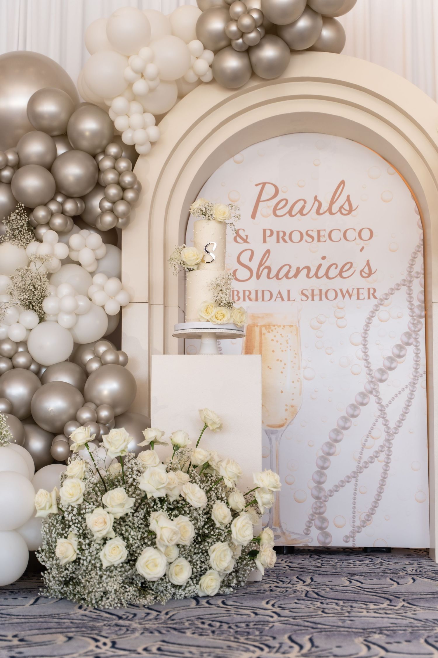 Elegant bridal shower backdrop with white and silver balloon arch and decorative white roses in front of arched panels.