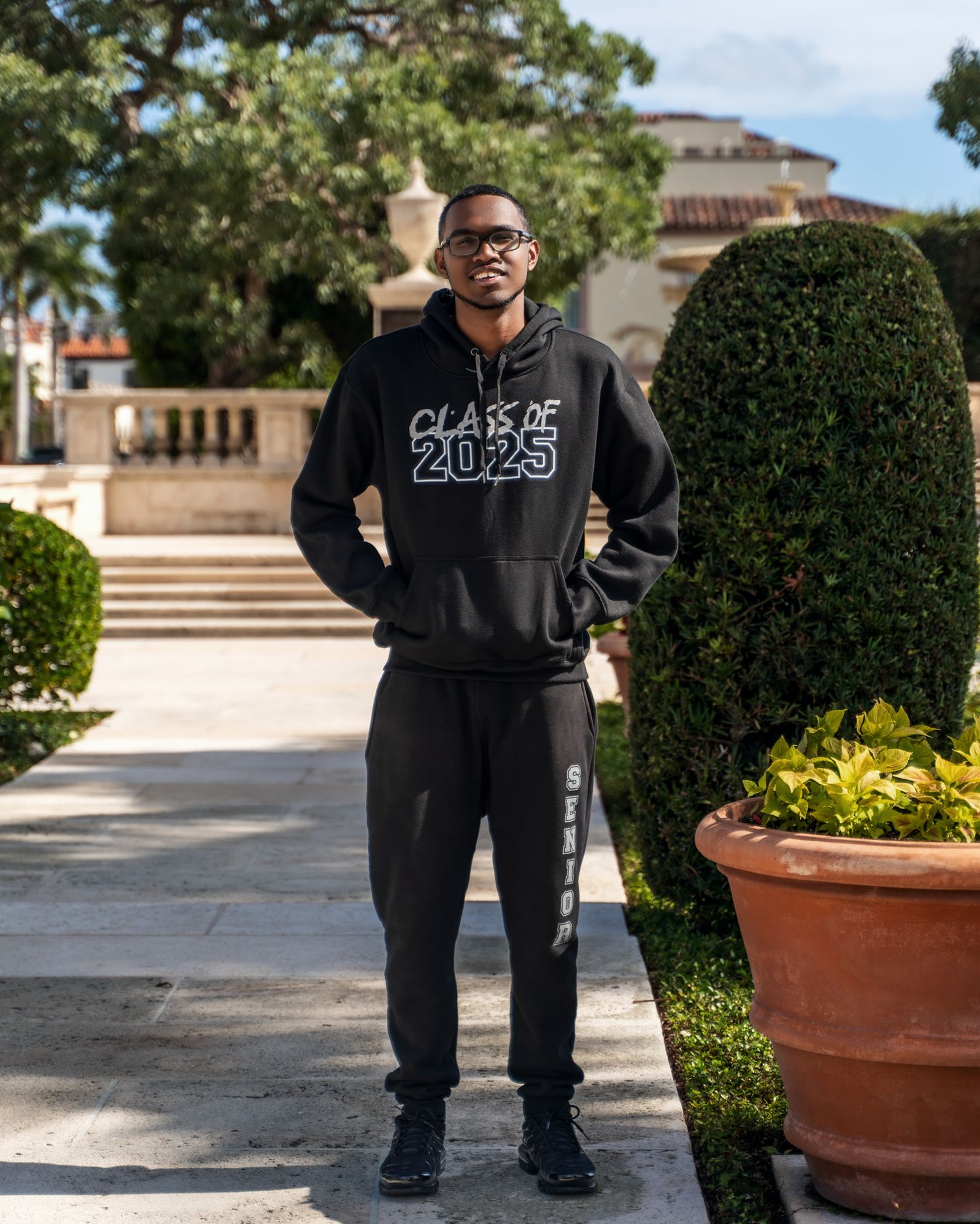 Student wearing black Class of 2023 hoodie and sweatpants stands on paved walkway with landscaped grounds and building in background.