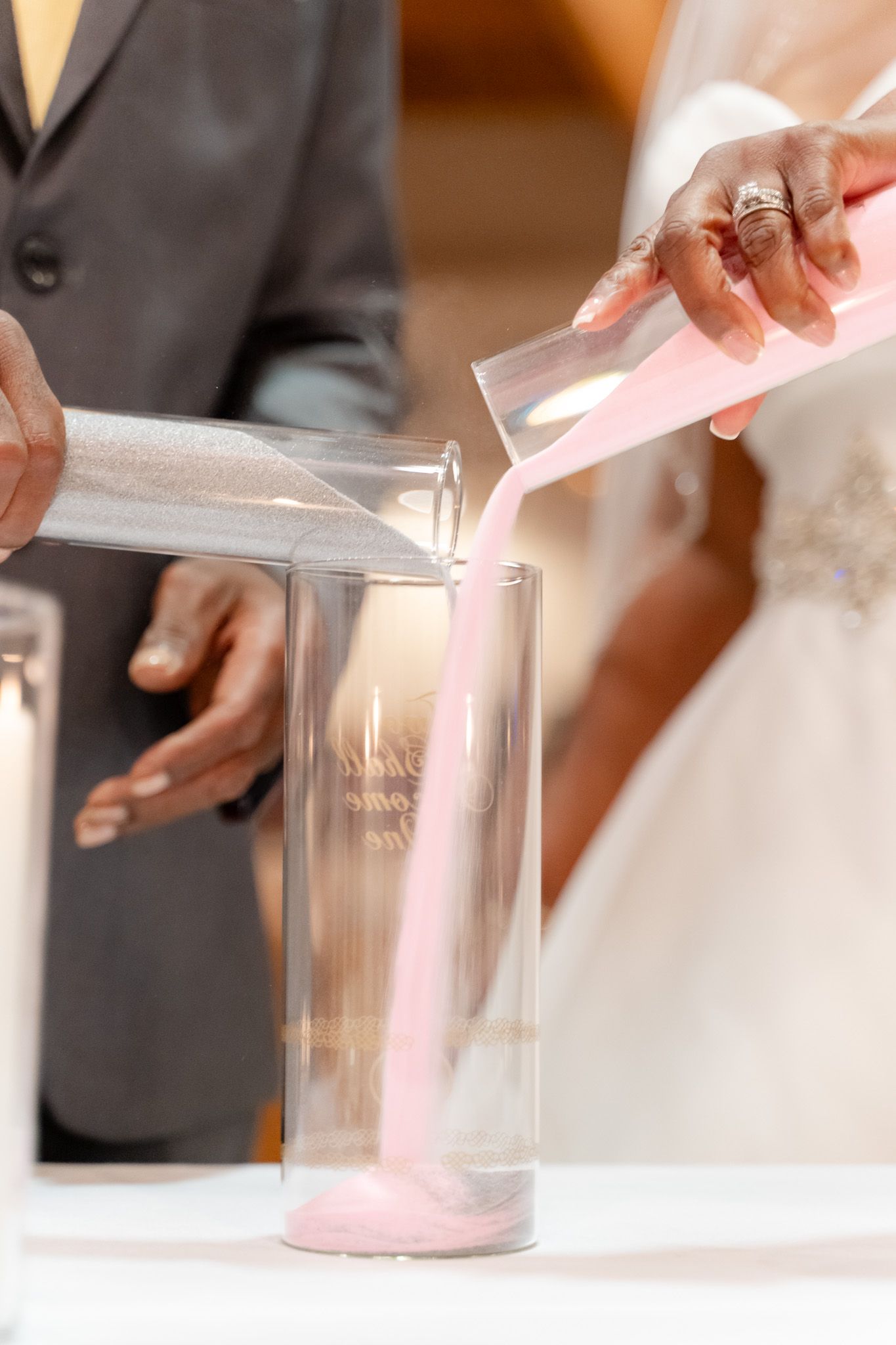 Couple performing unity sand ceremony pouring pink sand into glass vessel during wedding.