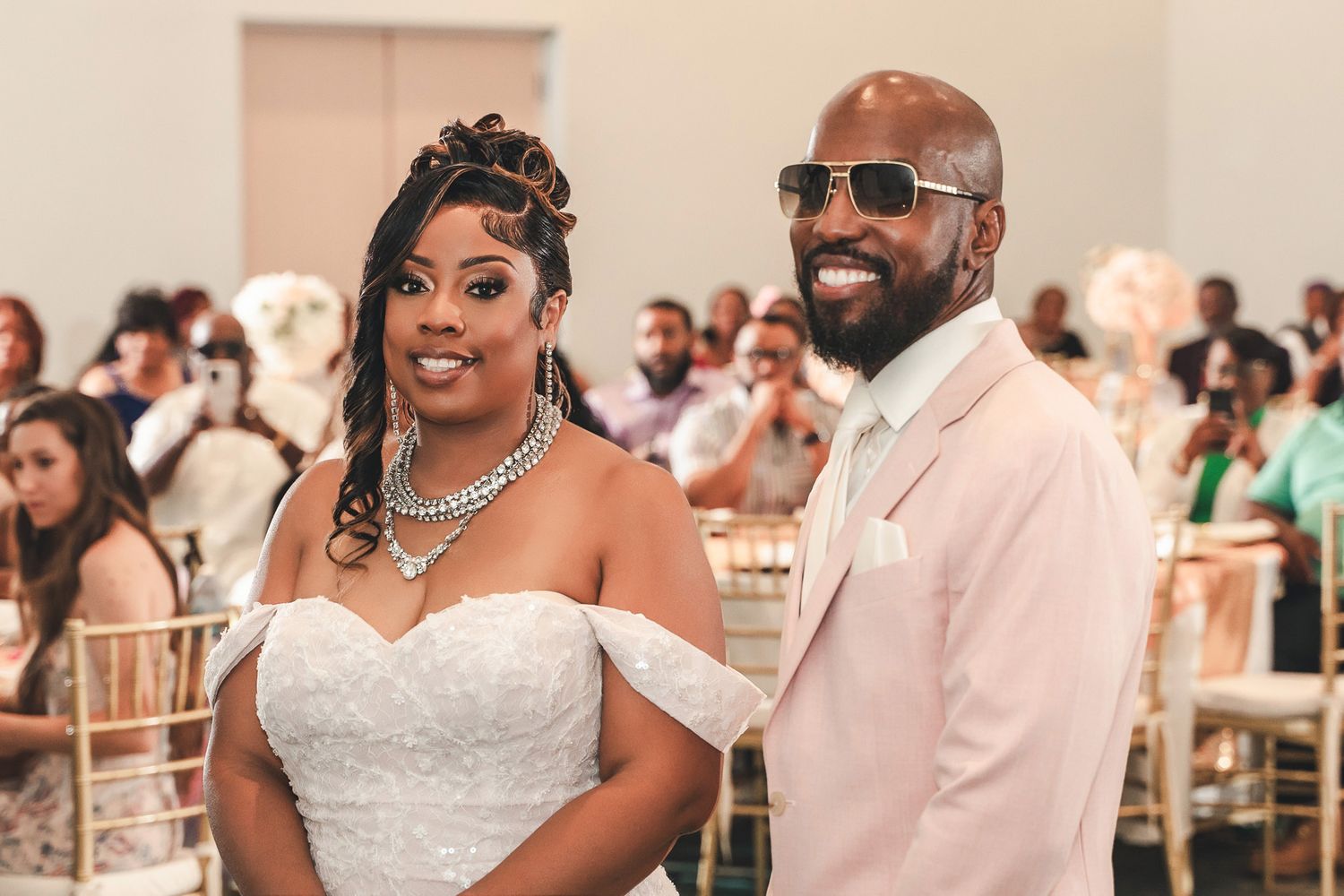A wedding couple in formal attire poses at their reception venue with the groom wearing a light pink suit and sunglasses.