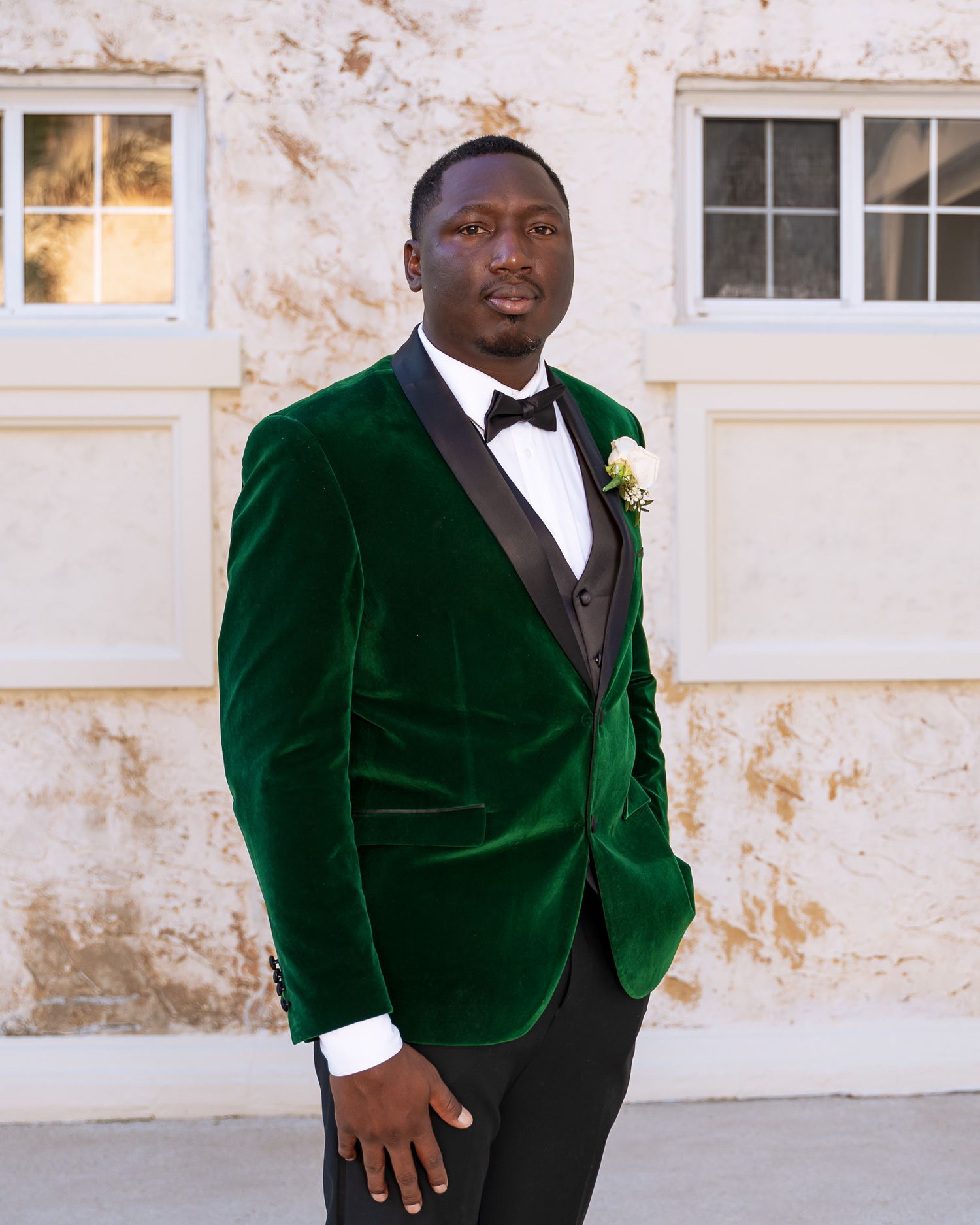 Groom in emerald velvet tuxedo poses elegantly against textured white wall.
