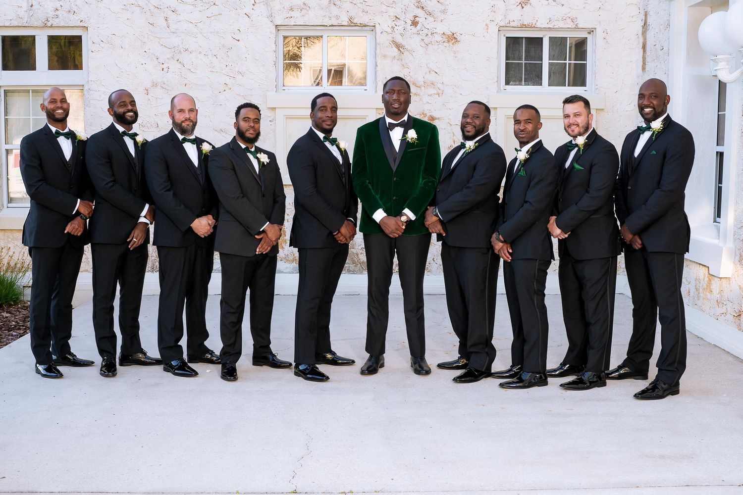 Groomsmen in black suits with groom in emerald velvet jacket pose against white wall for formal portrait.