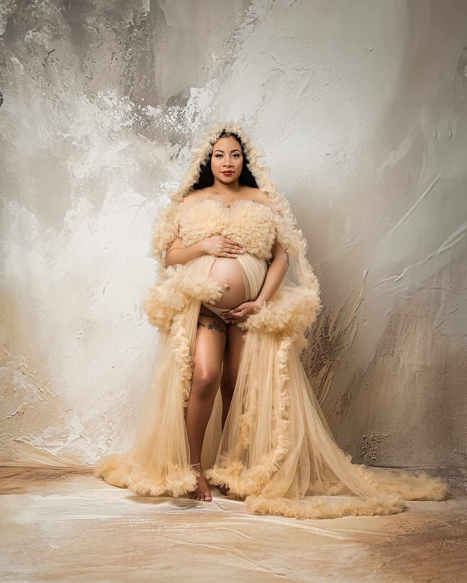 Ethereal maternity photo featuring flowing champagne tulle gown against textured beige wall.
