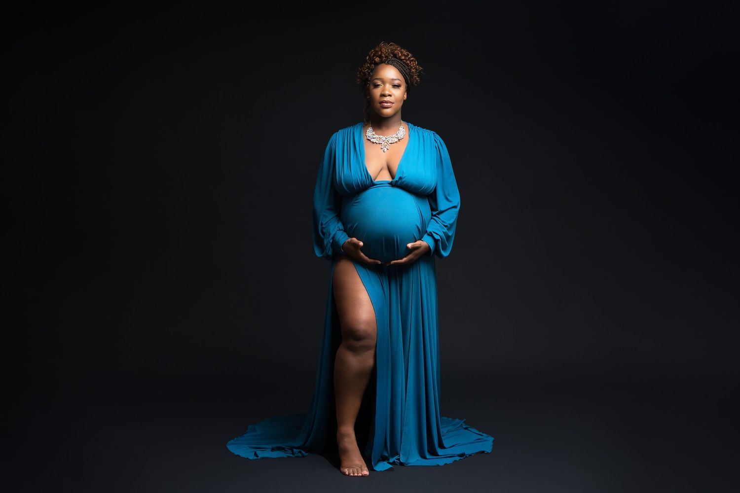 A regal maternity portrait features an expectant mother in a flowing blue gown against black background.