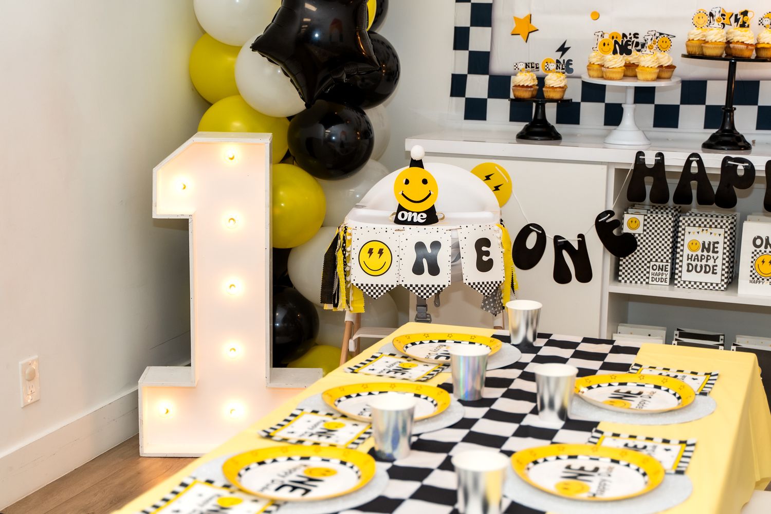 First birthday party decorations with large light-up number one, yellow and black balloons, and smiley face theme.