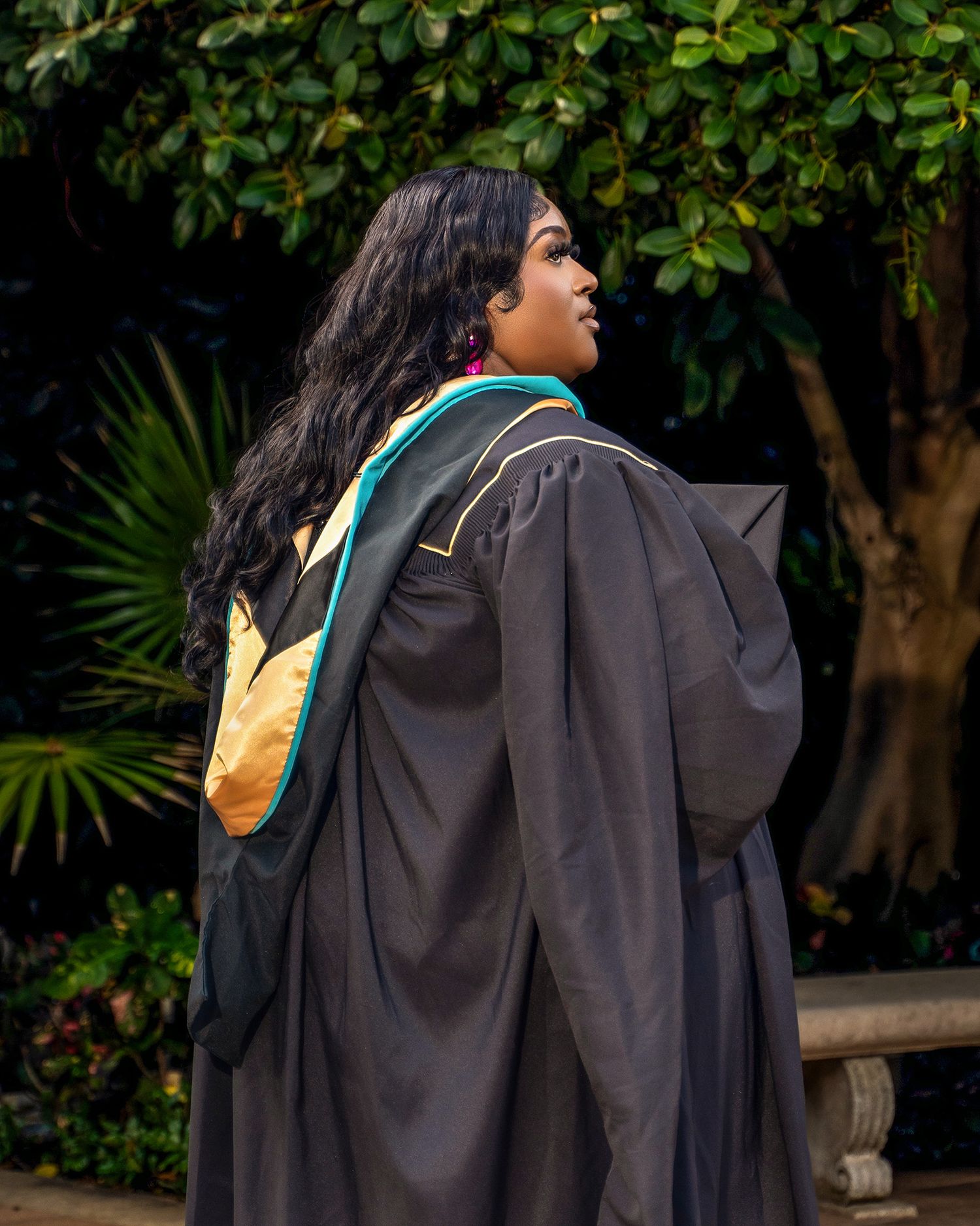 Graduate in academic regalia poses in front of lush garden backdrop.