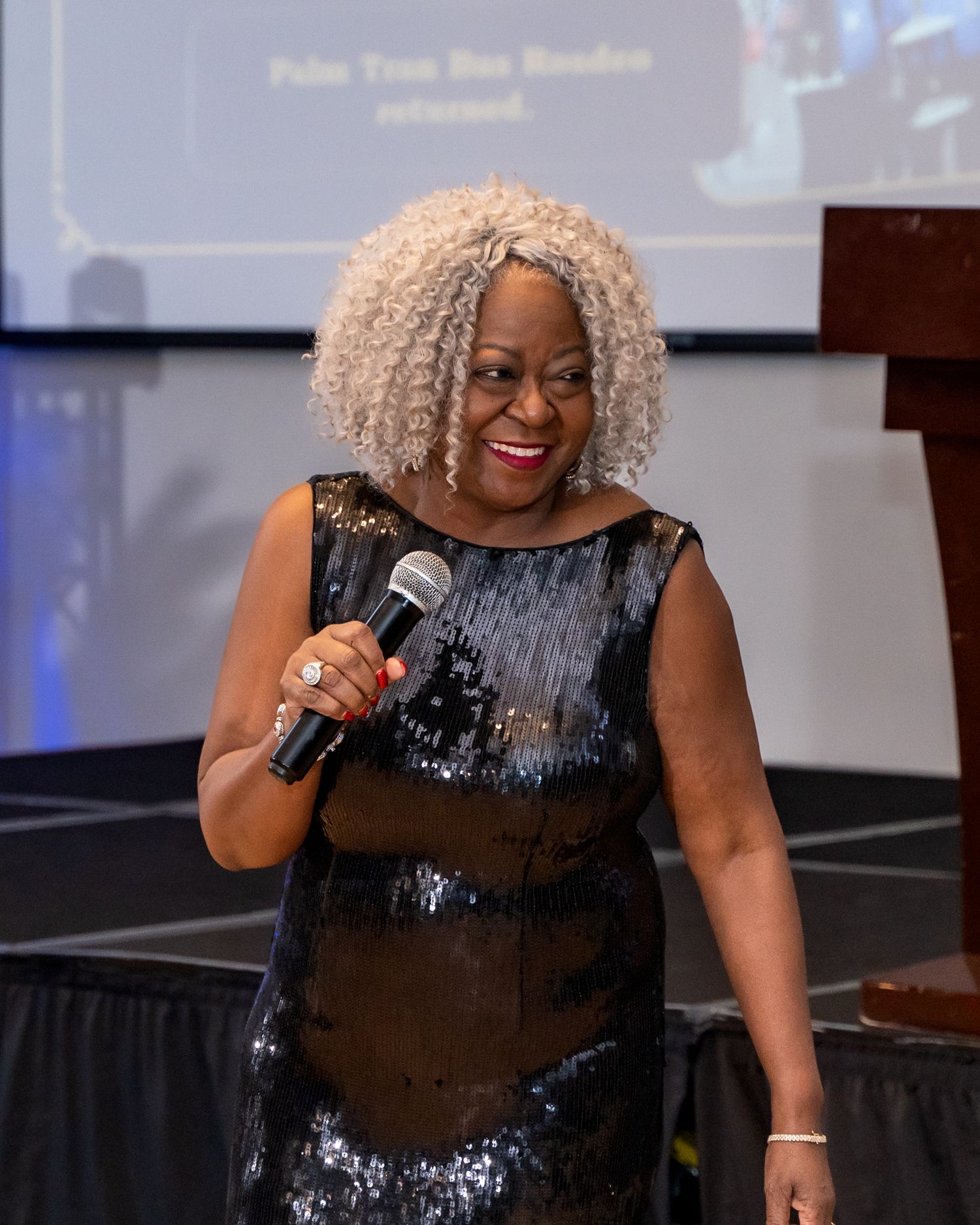 A performer in a sparkly black dress holds a microphone on stage during an event.