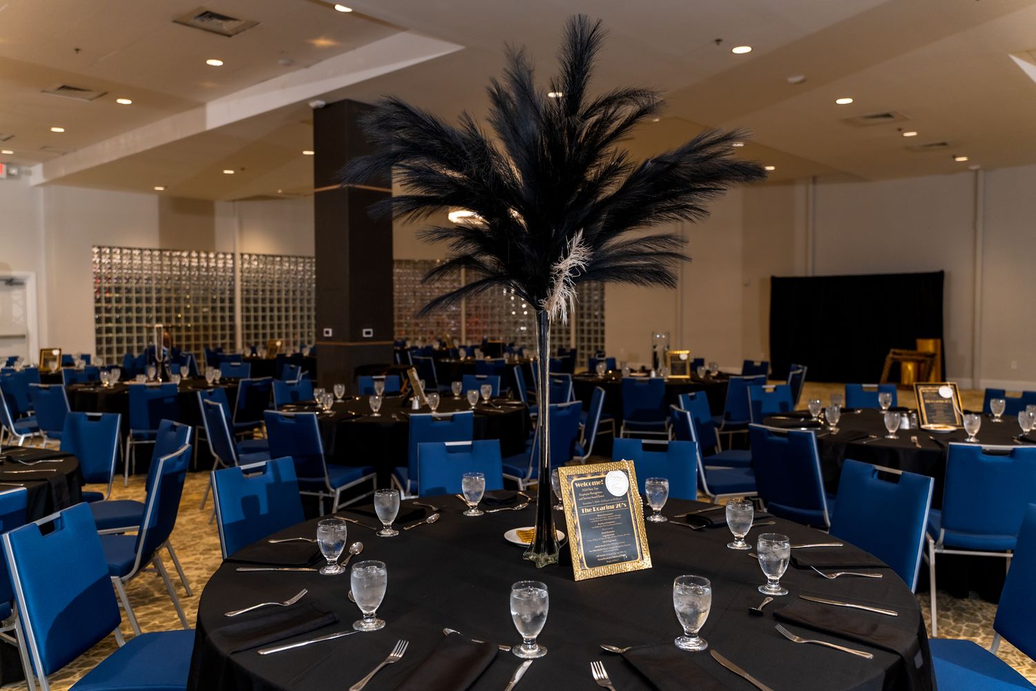 An elegant banquet hall setup with blue chairs and black tablecloths centered by tall black feather centerpieces.