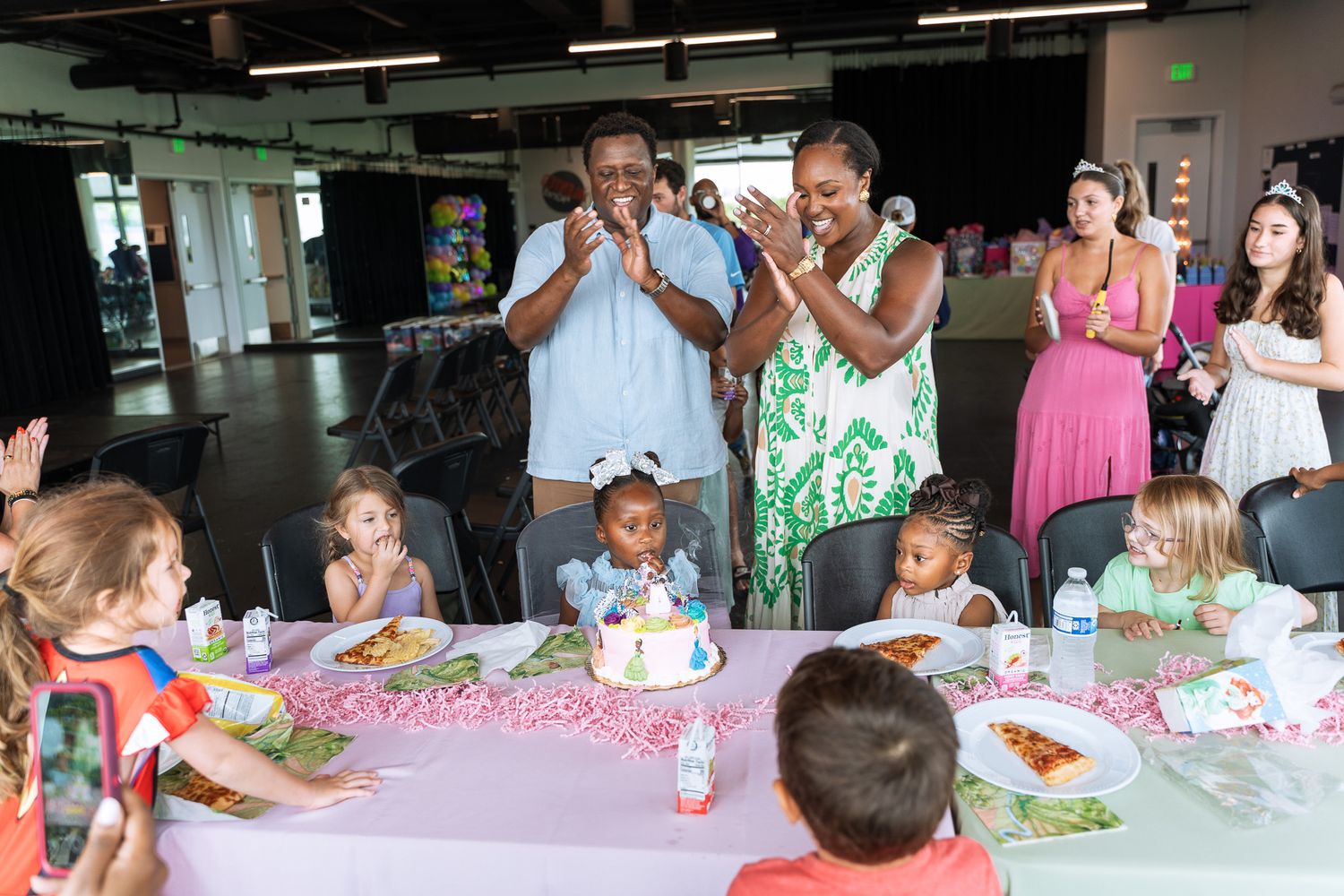 A joyful celebration around a birthday cake with guests clapping and singing at a party.