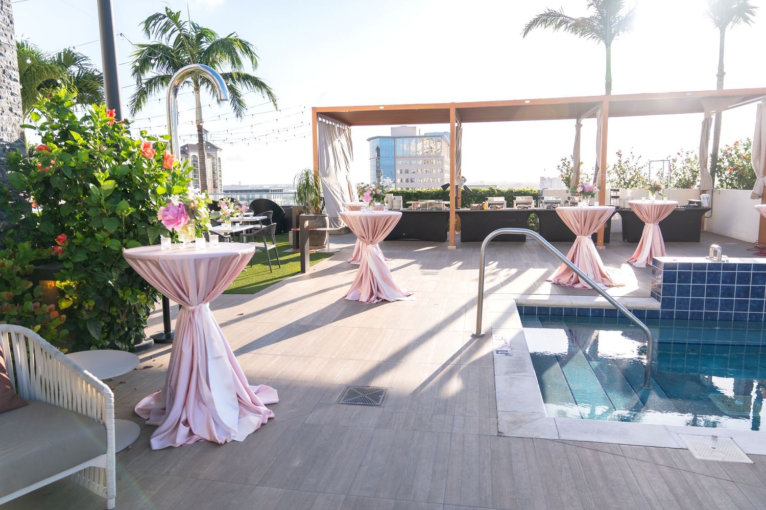 Elegant outdoor event space with pink cocktail tables and a small pool on a rooftop terrace surrounded by palm trees.