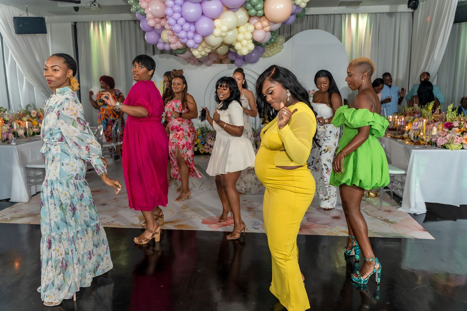 A group dances together at an event decorated with pastel balloon arches and floral arrangements.