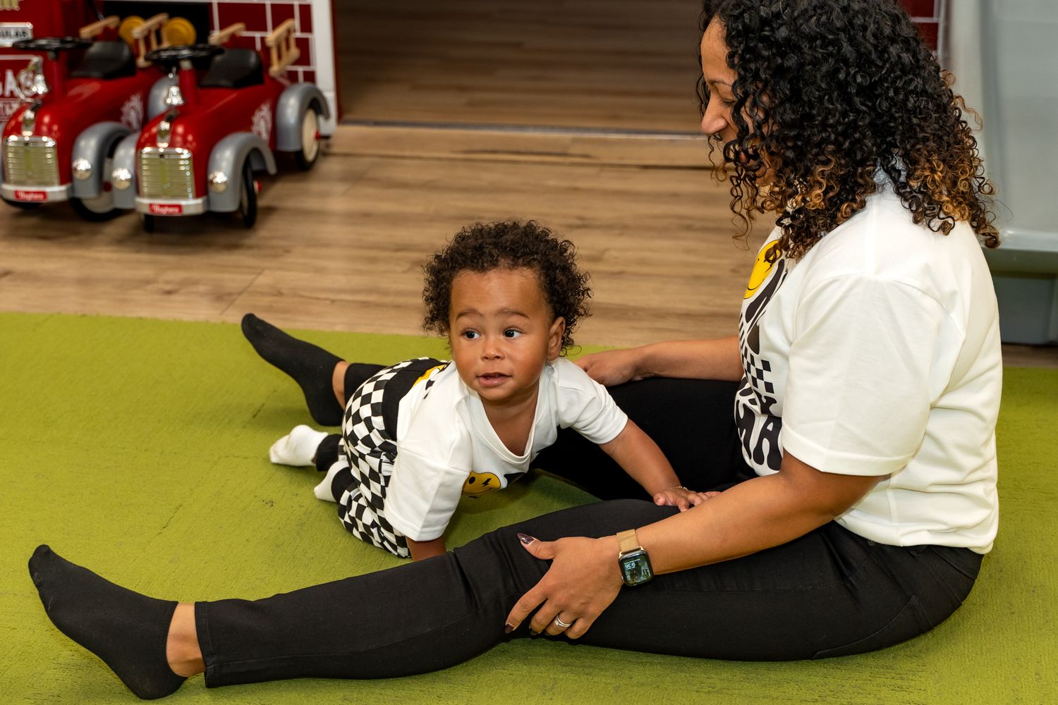 Child playing on green carpet near toy fire trucks while caregiver watches.