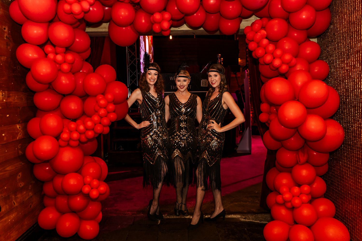 Three people in 1920s flapper costumes posing under a red balloon arch.