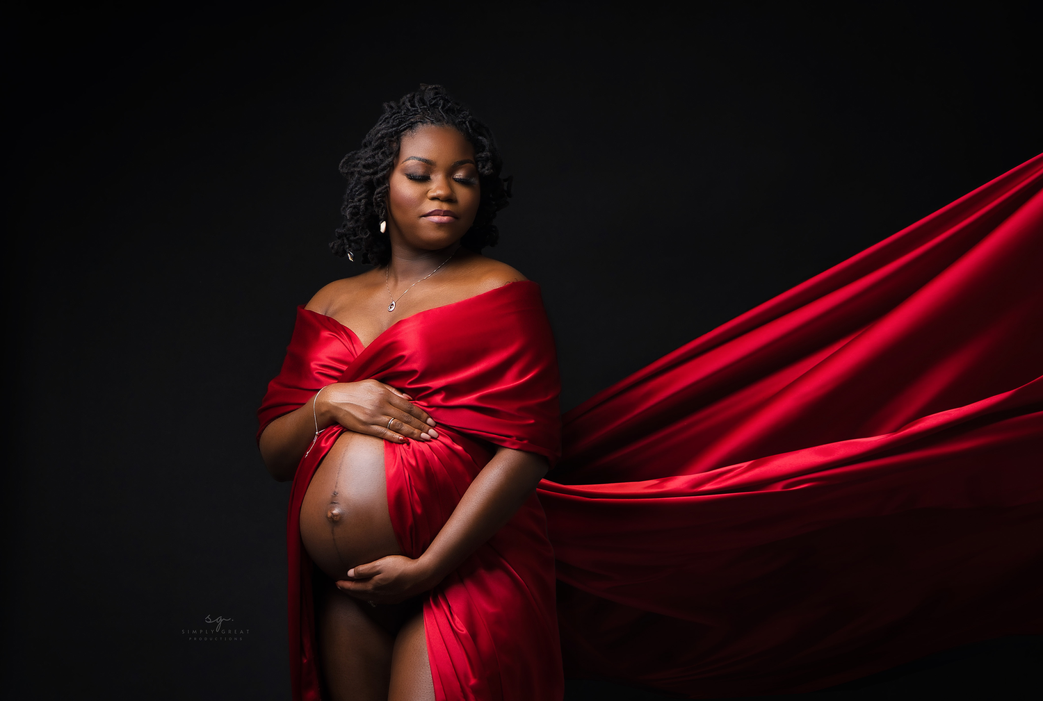 Elegant maternity photo with billowing red satin dress against black backdrop.